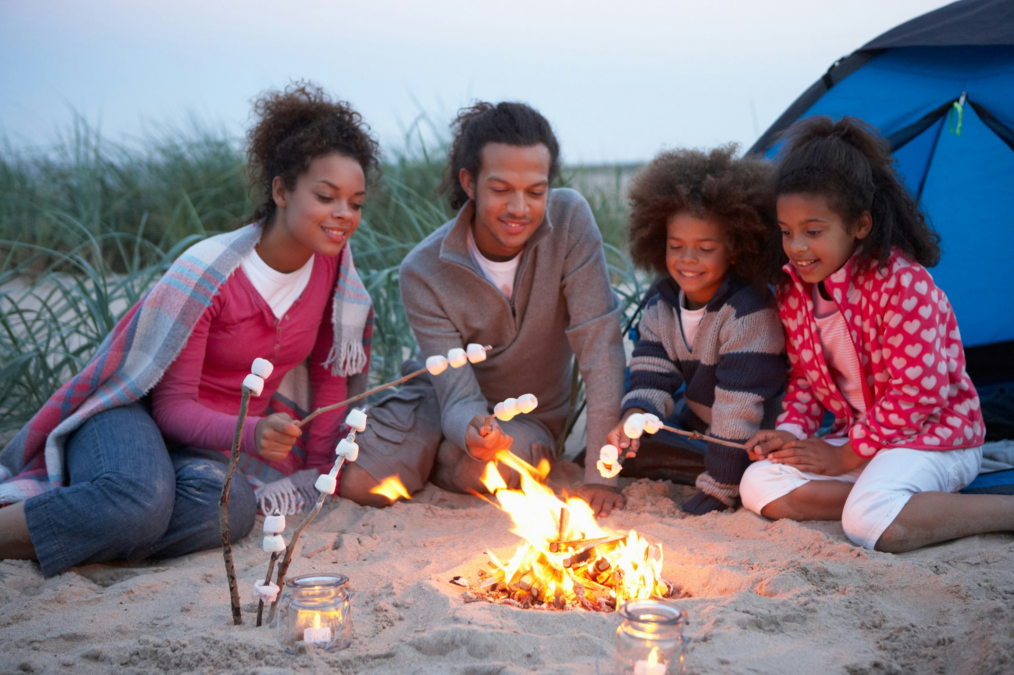 A family of four roast marshmallows by their tent on the beach.