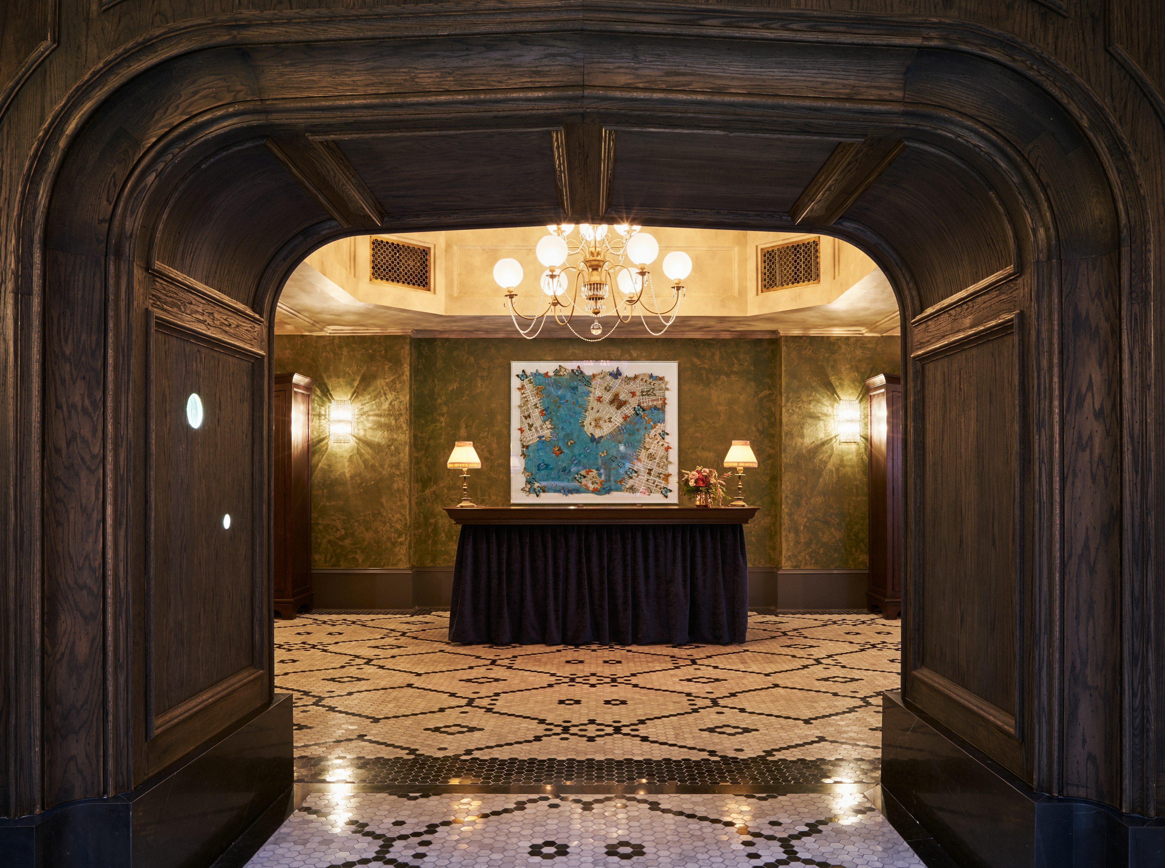 A grand, dark-wood archway leads to a room with an ornately tiled floor and a concierge desk under a chandelier at The Beekman. Jane Hammond’s 'All Souls (Buttermilk Channel)' (2015) is hanging on the wall behind the desk.