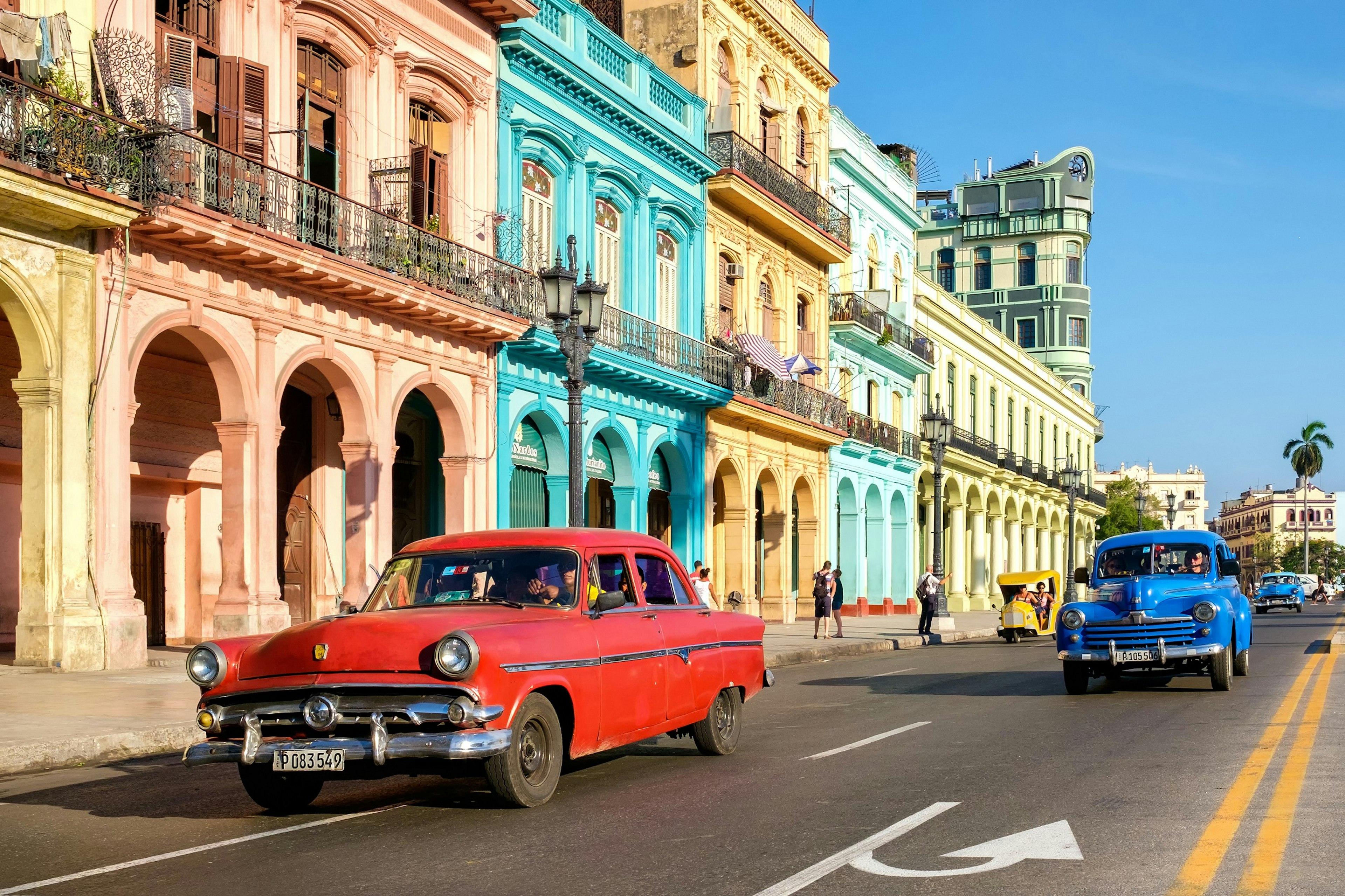 A run-down looking classic American car drives down a street in Havana; the buildings are well preserved and vibrantly colored.