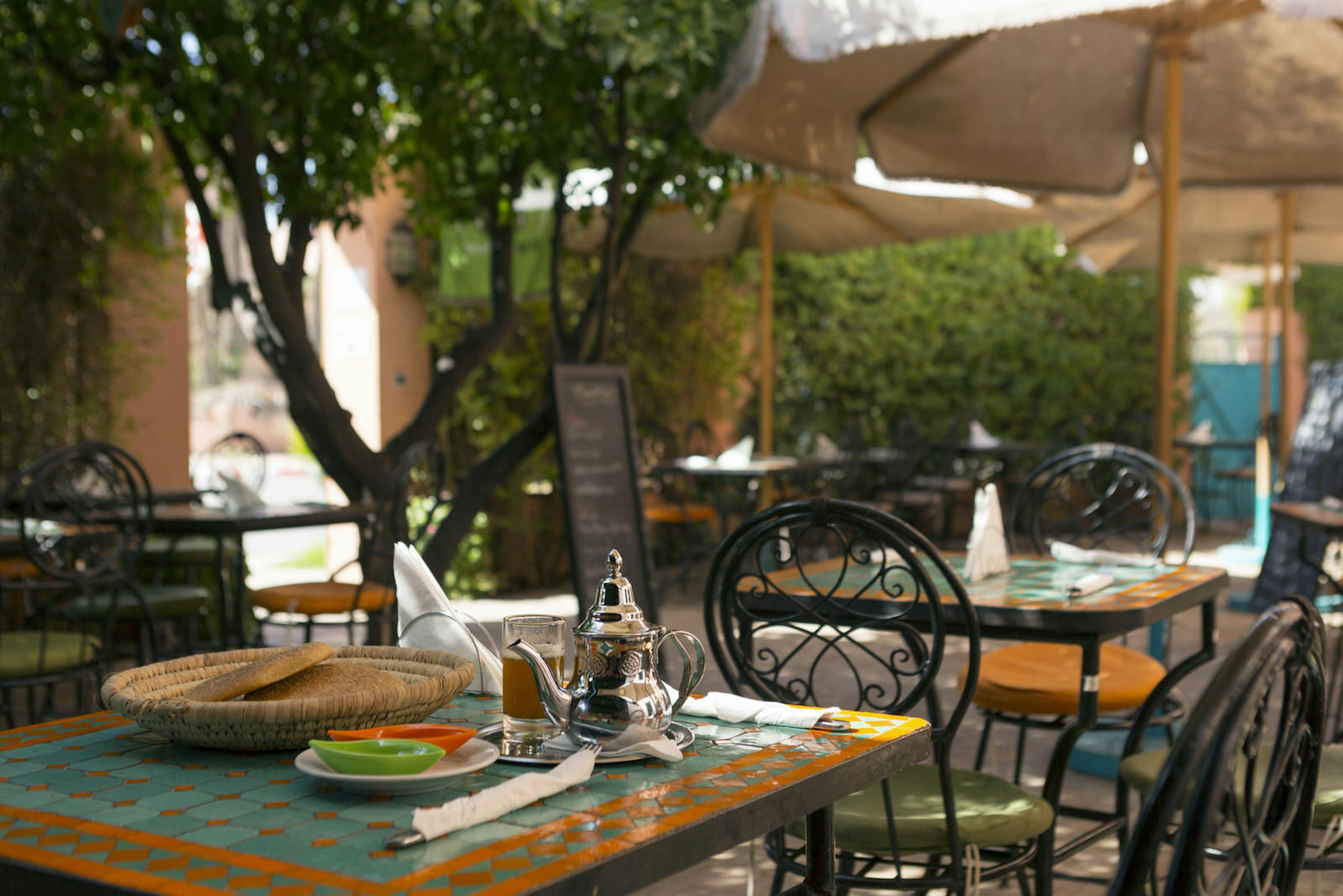 Mint tea in an ornate silver teapot and bread in a straw basket on a colourful, tiled table in the cafe courtyard at Amal, Marrakesh, Morocco. The tables are shaded by leafy trees and white umbrellas and have cast-iron chairs to sit on.