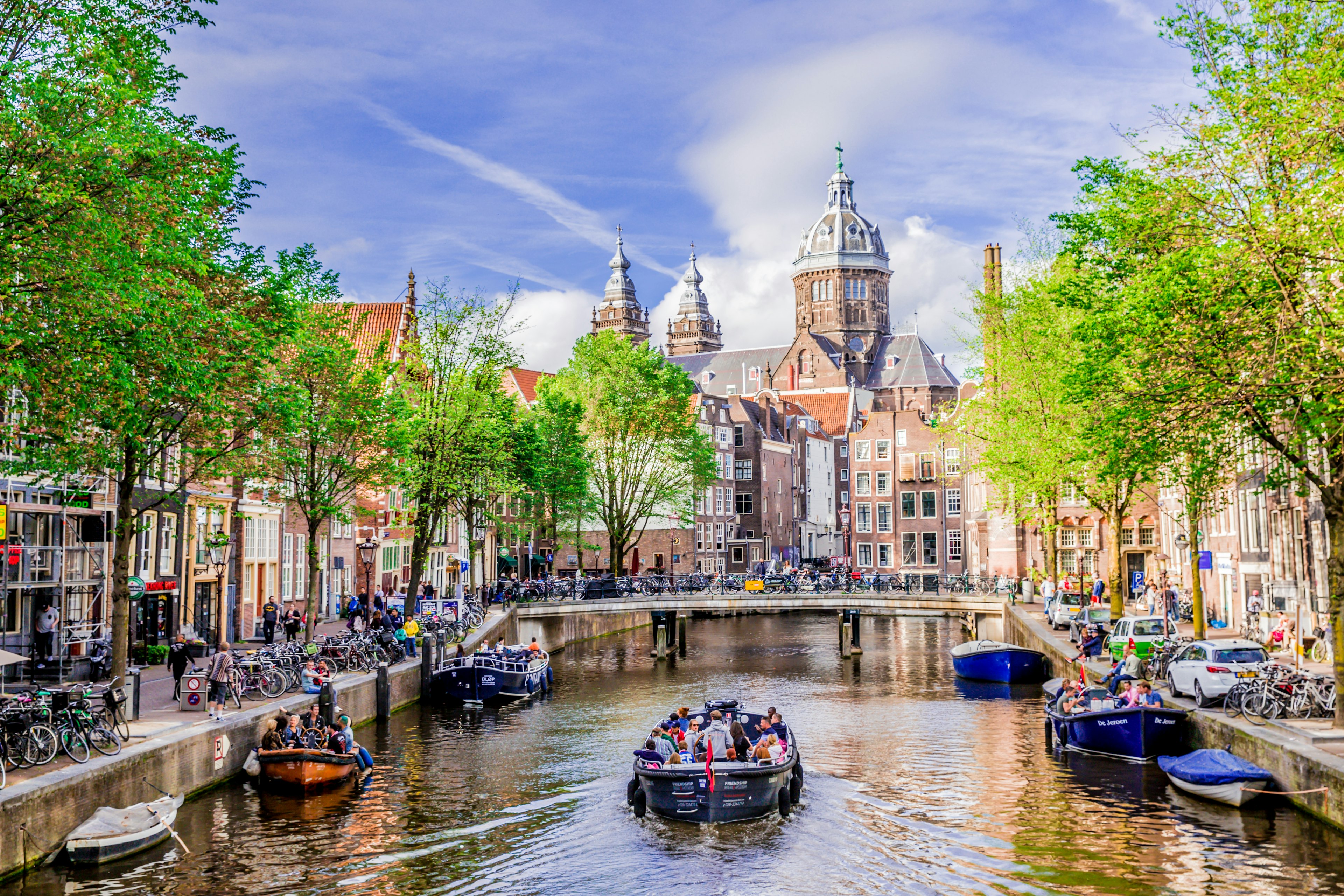 A boat tour is gliding down one of Amsterdam's canals on a sunny day. Several boats line the edge of the canal and the streets are busy with bikes and people.