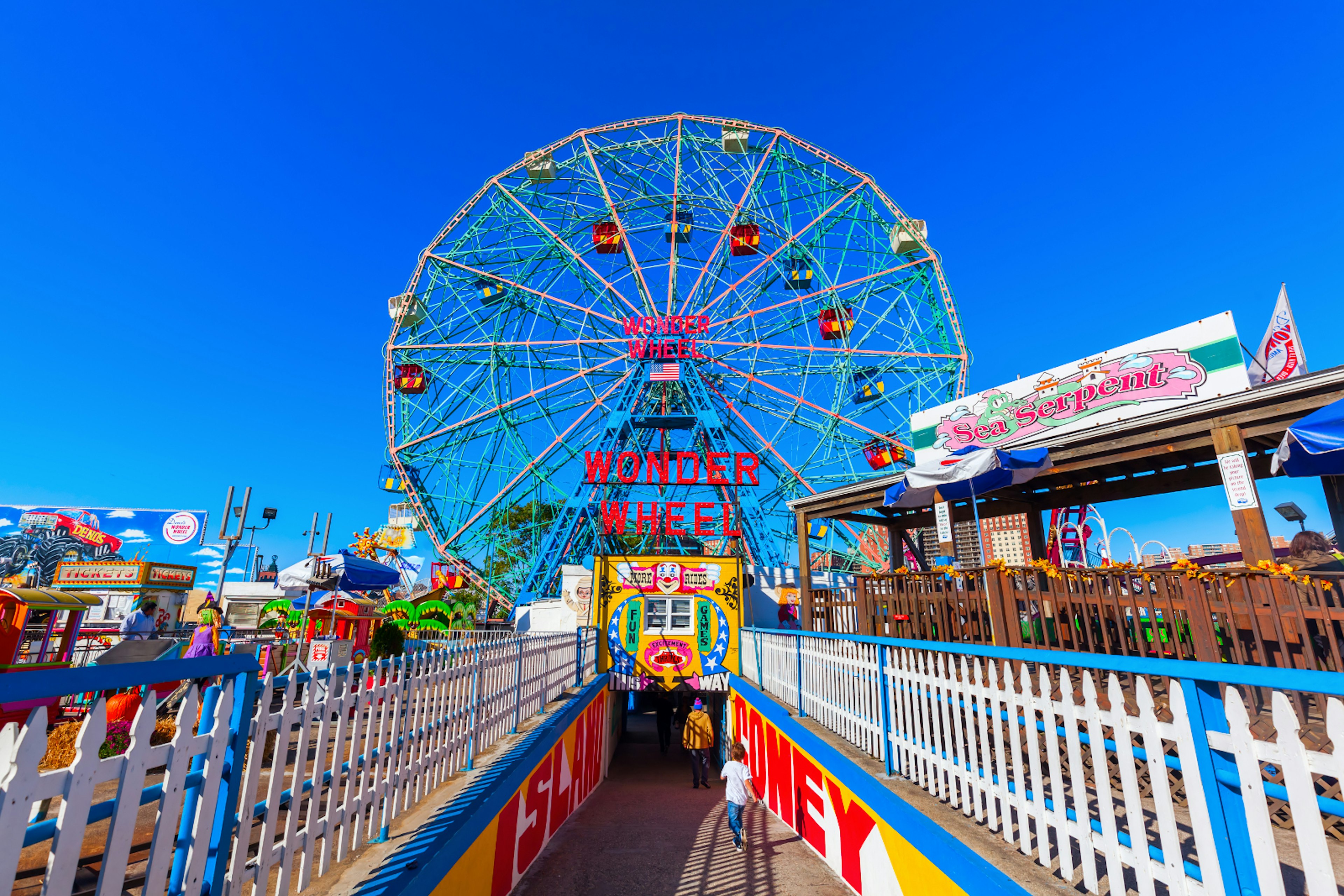 A colourful ferris wheel on Coney Island stands in front of a brilliant blue sky, with colourful railings and placards flanking the tunnel down to the wheel's entrace