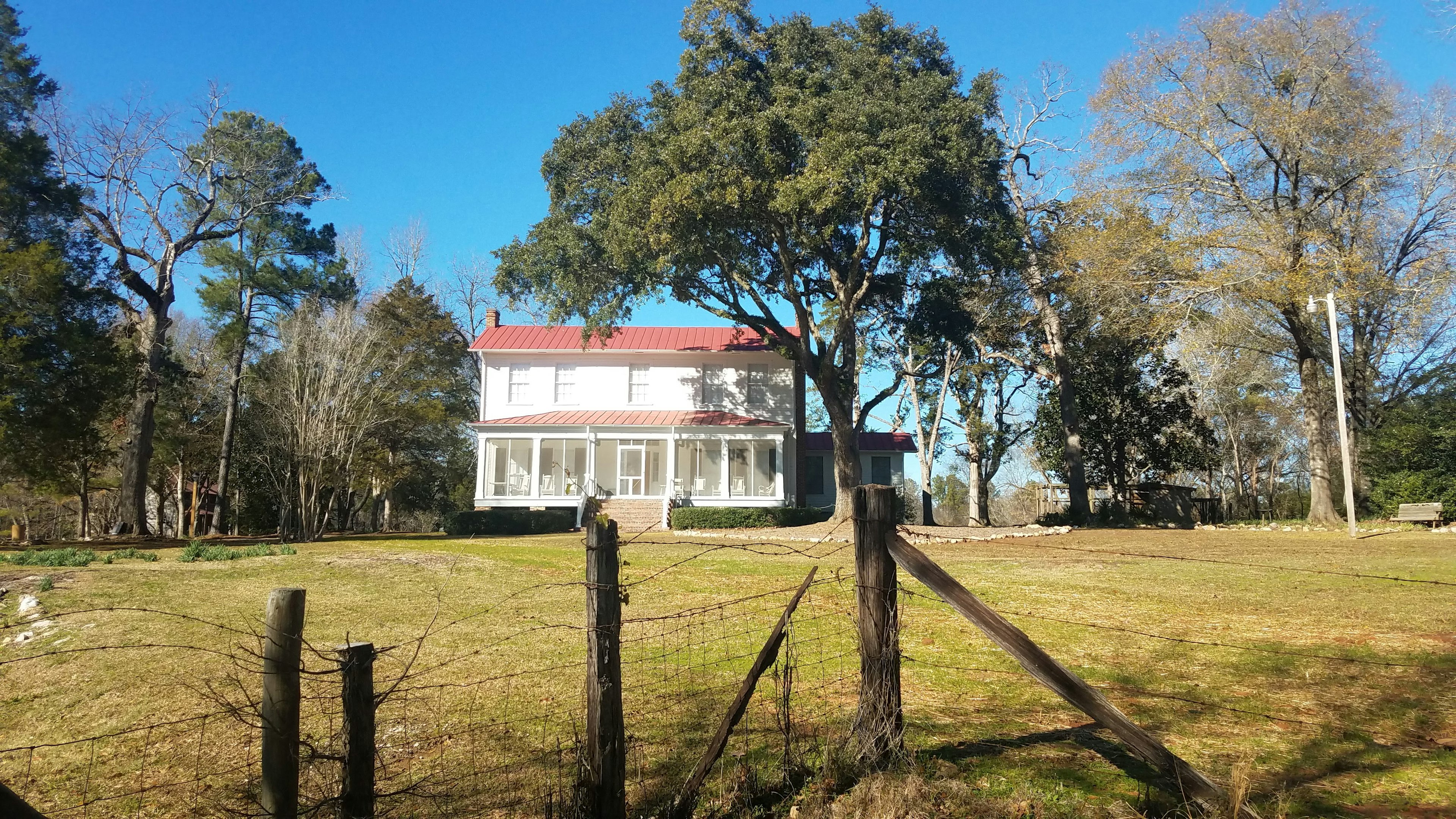 The white clapboard exterior of Flannery O'Connor's Milledgeville, Georgia farmhouse with a red roof sits beneat hteh shade of a massive old tree. A large green yard spreads out in front of the house up to an old wire fence in the foreground.