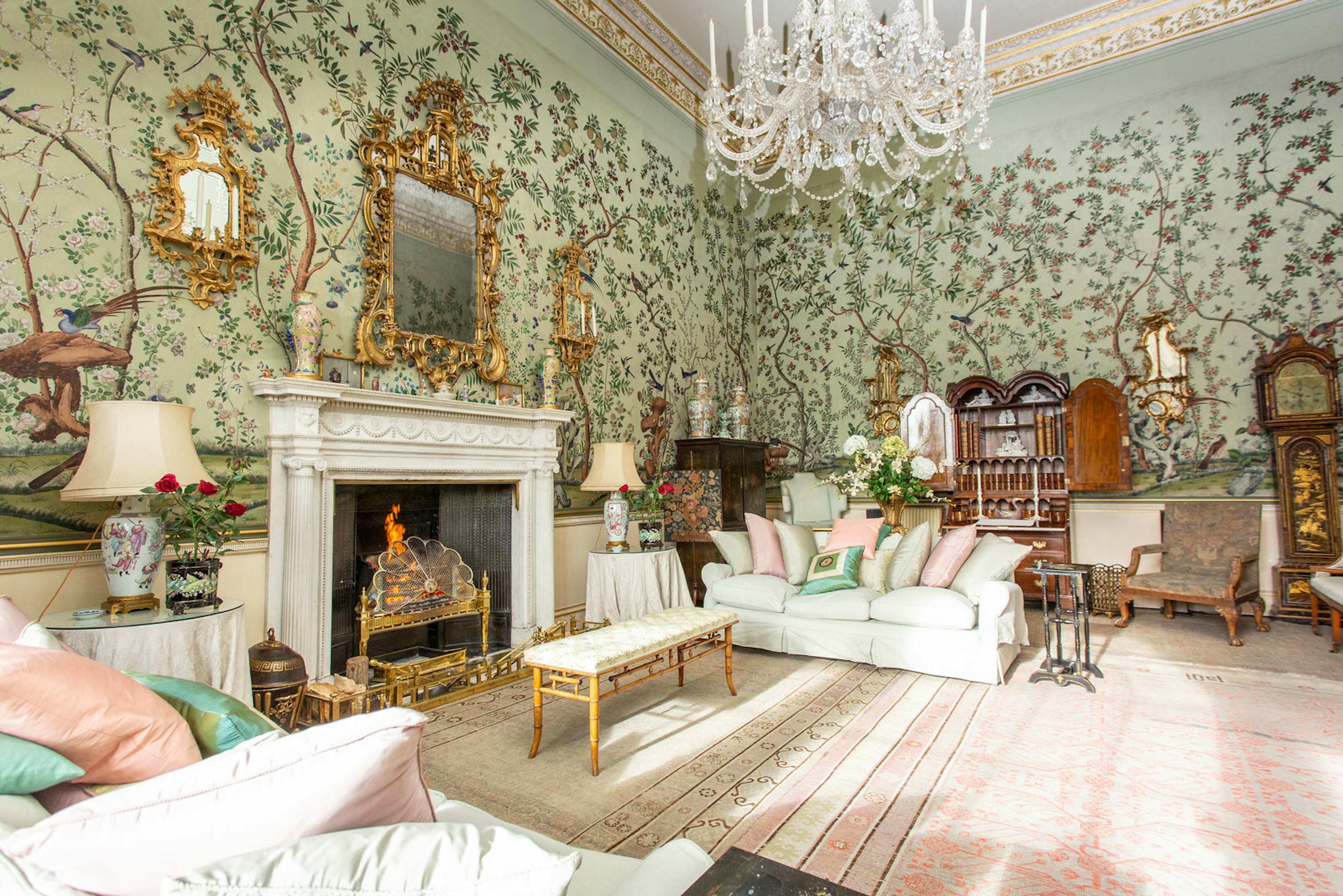 A Georgian-era room decorated in opulent style with pink brocade carpet, marble fireplace, chandelier and gold framed mirror.