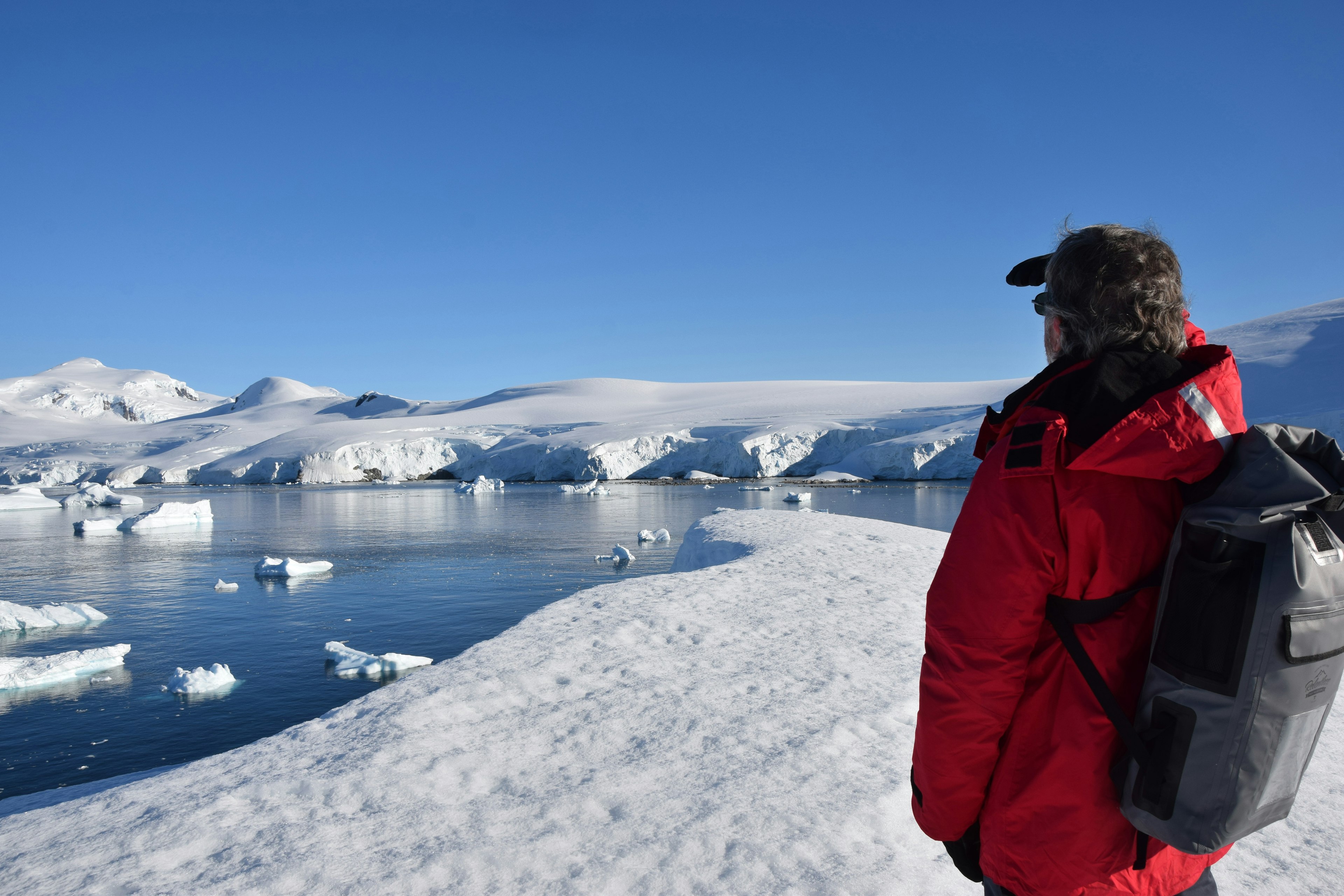 Michael Ballard stands with his back to the camera in a red cruise-line parka and grey dry-bag style backpack gazing out across the snow and ice fields of Antarctica