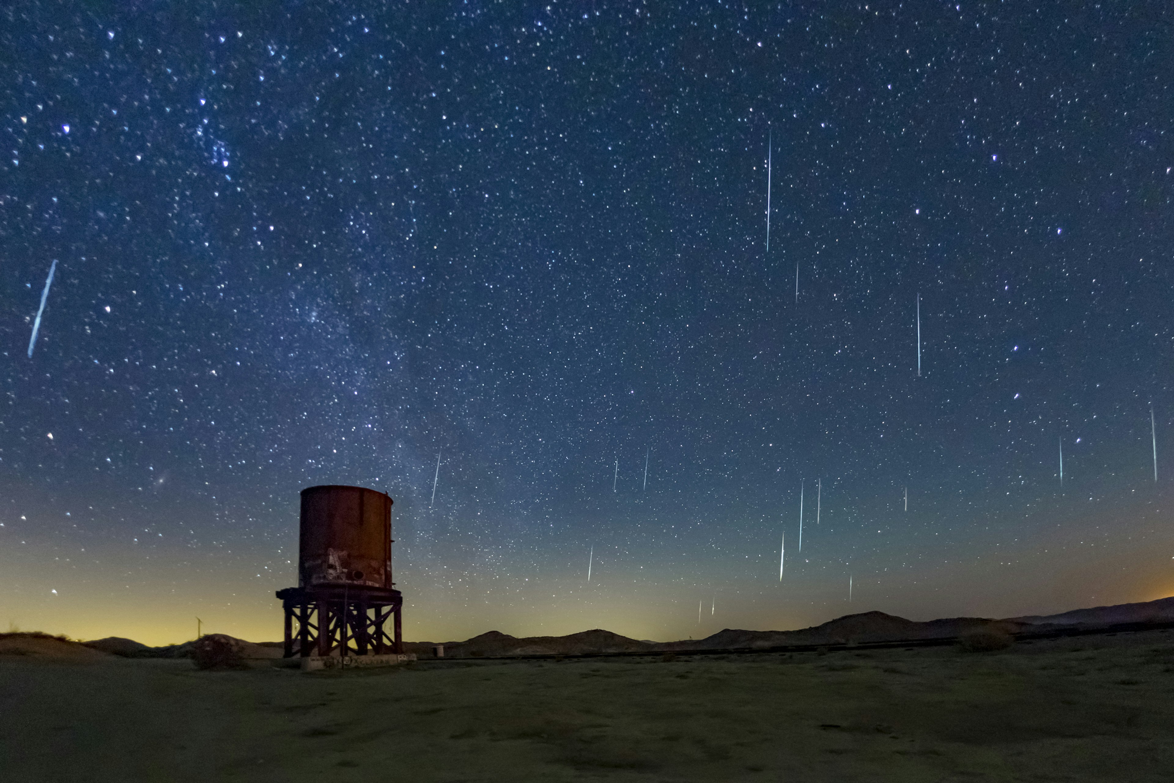 The Geminids Meteor Show in 2017 falls over Anza Borrego Desert State Park. The sky is a bright, warm indigo blue dotted with white and light blue stars and streaks from the falling meteors. The horizon is a light yellow or deep orange and framed by low mountains and hills. In the mid ground is a red water tower, part of the old Dos Cabezas Siding