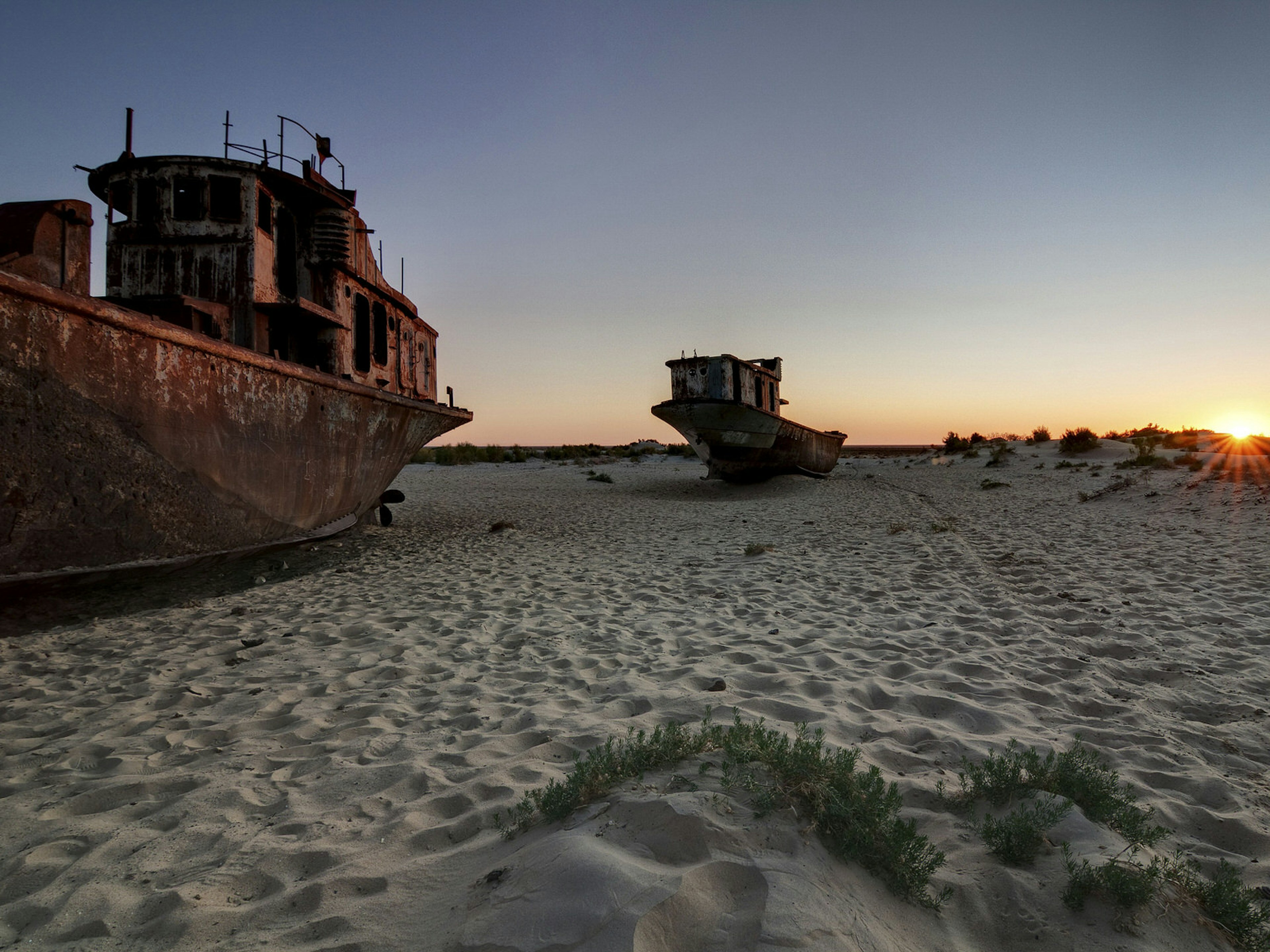 Rusting ships beached on sand with the sunset behind.