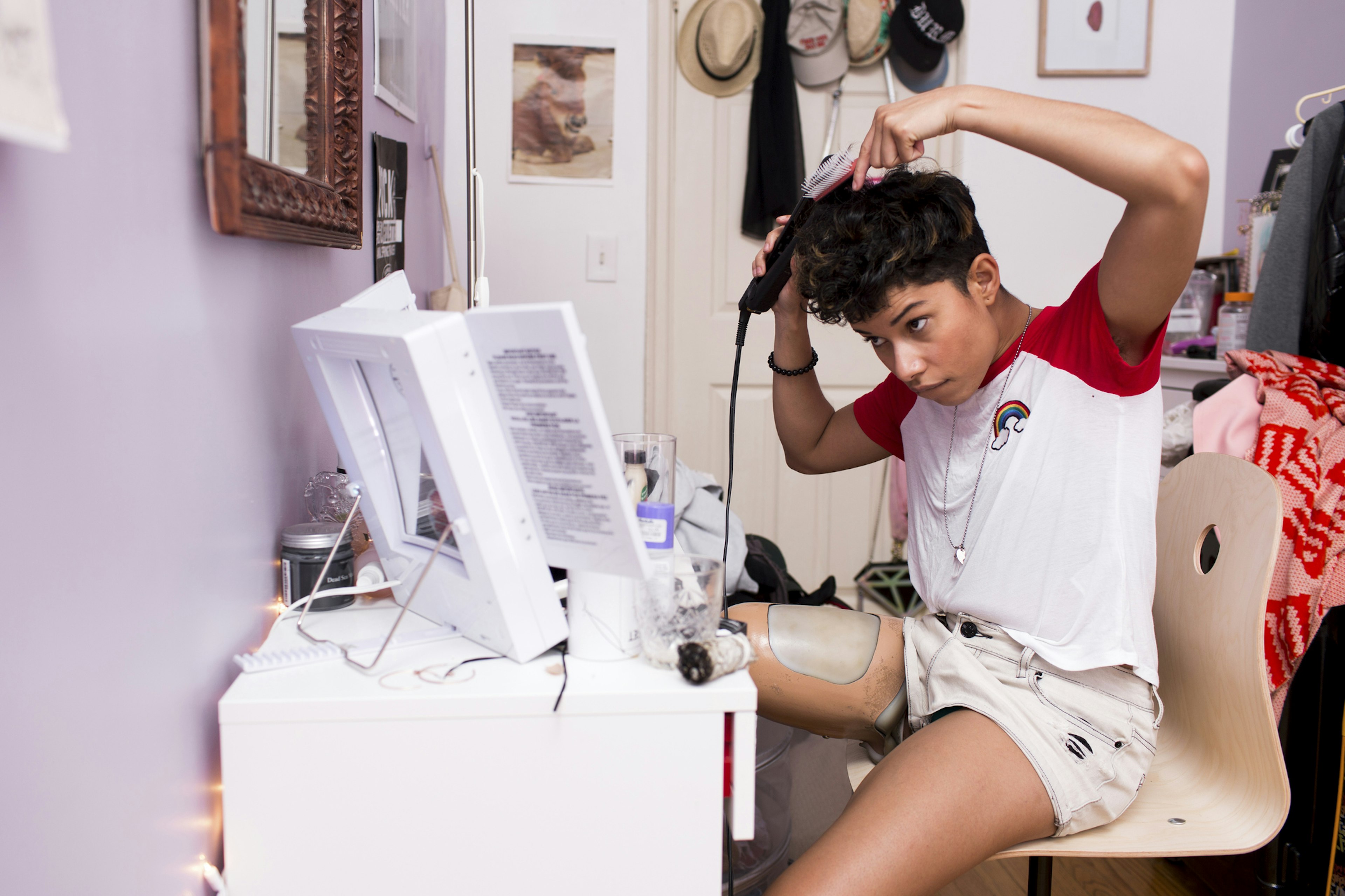 A young woman with prosthetic right leg styles her short hair in front of a vanity table with a large light-up mirror