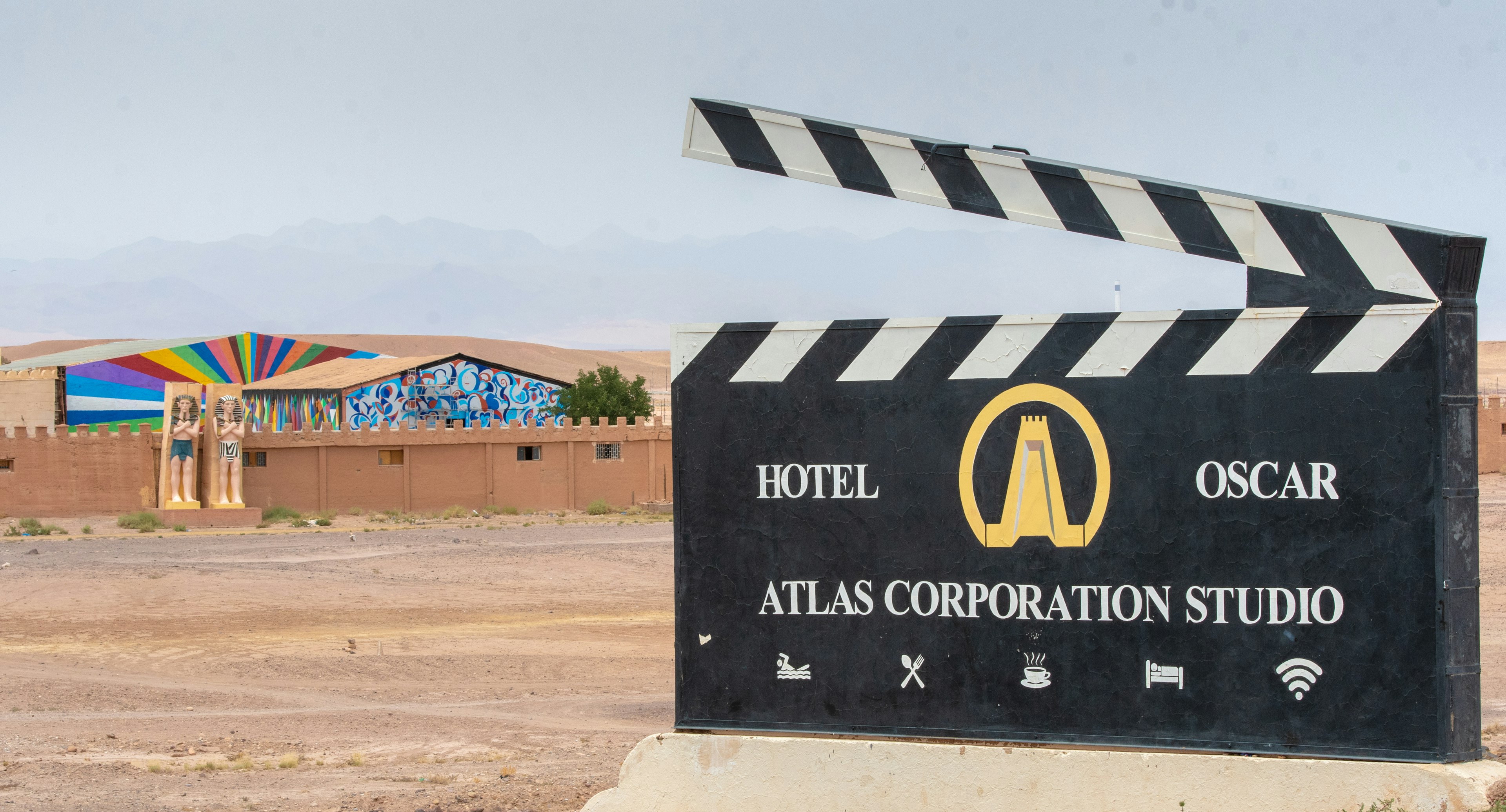 The entrance to Atlas Studios in Morocco looks like a large black clap board with a stylized illustration of a golden tower like those seen at nearby Ait Ben Haddou. Behind the sign is a crenelated wall, Egyptian style statues, and a rainbow sunburst mural