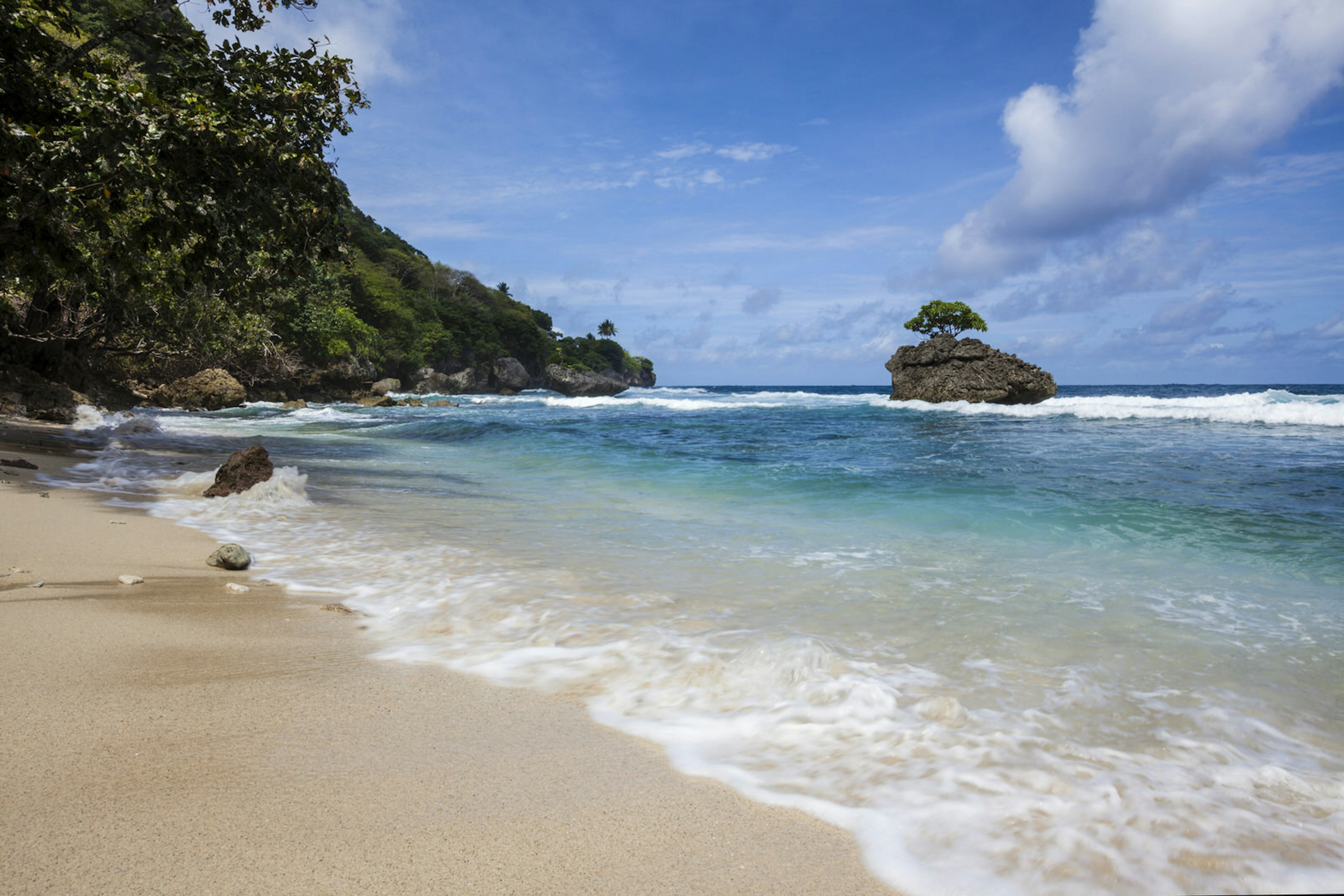 Waves lap against a golden sand beach and a boulder-covered, forested shore in the distance