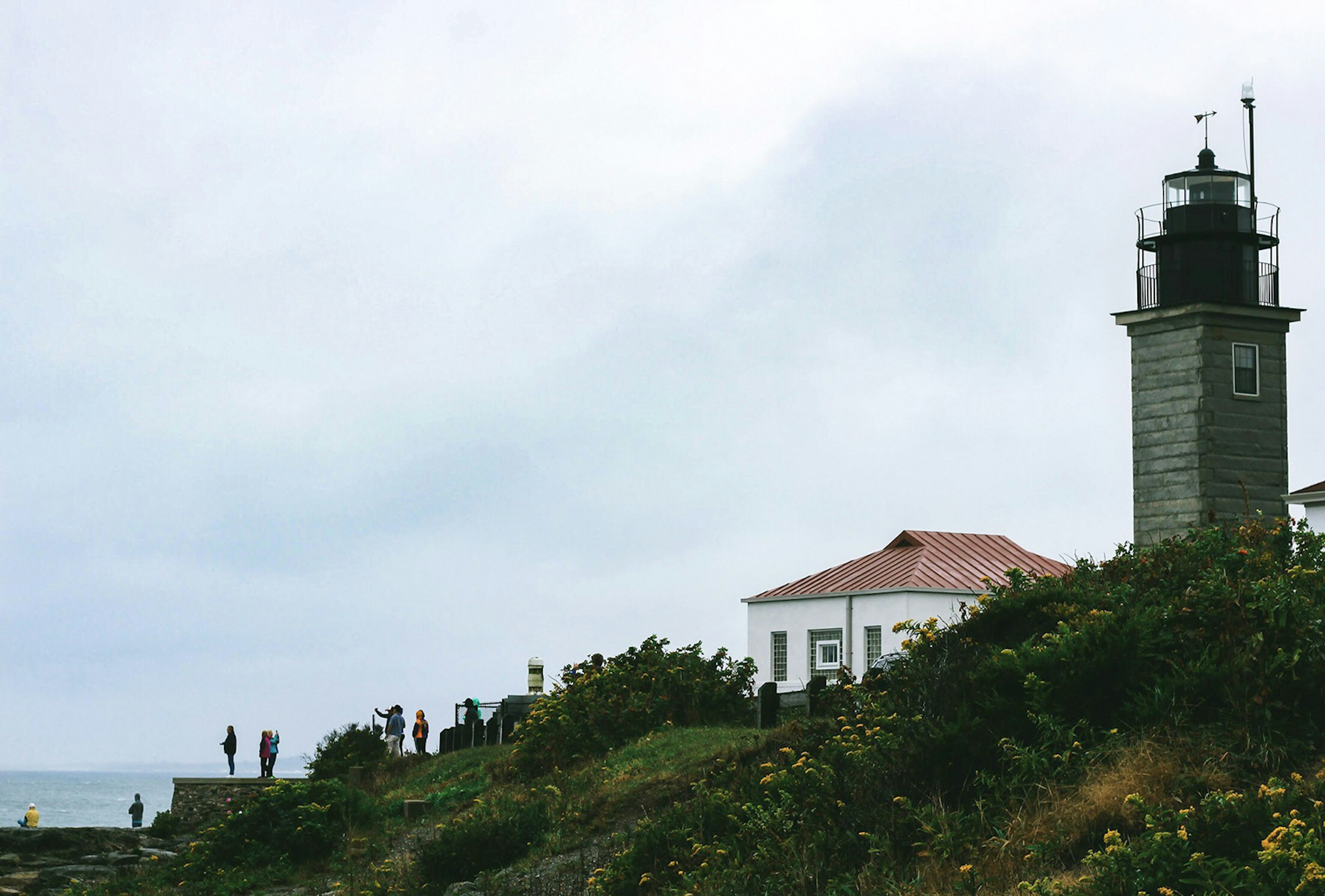 Distance view of Beavertail Lighthouse and the keeper's house, from a side angle. Shoreline is green and rugged, and the sky is overcast © Anna Saxon / Lonely Planet