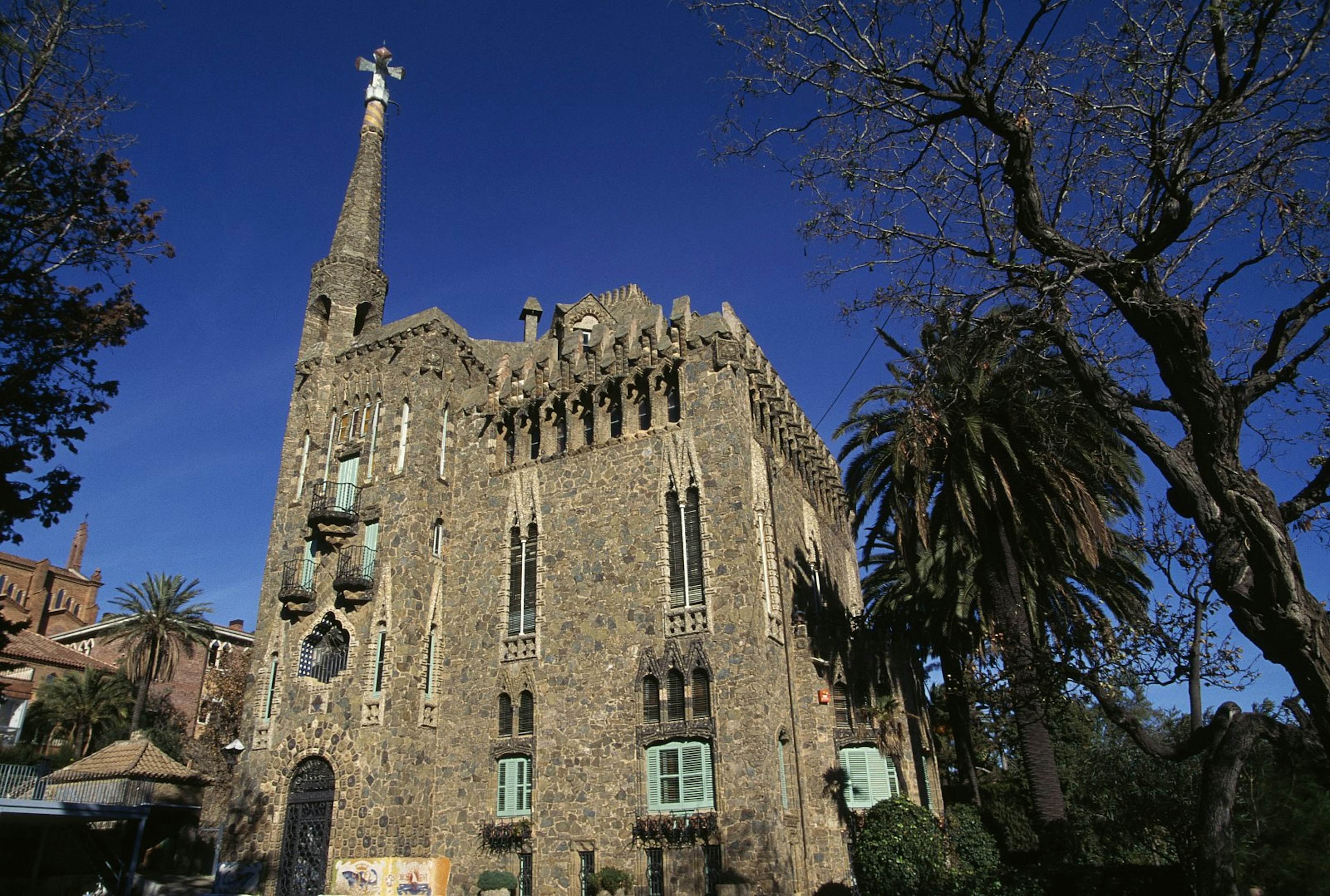 Bellesguard: it has a castle-like appearance with crenellated walls of stone and brick, narrow stained-glass windows, elaborate ironwork and a soaring turret mounted by a Gaudían cross. There are trees in its grounds.