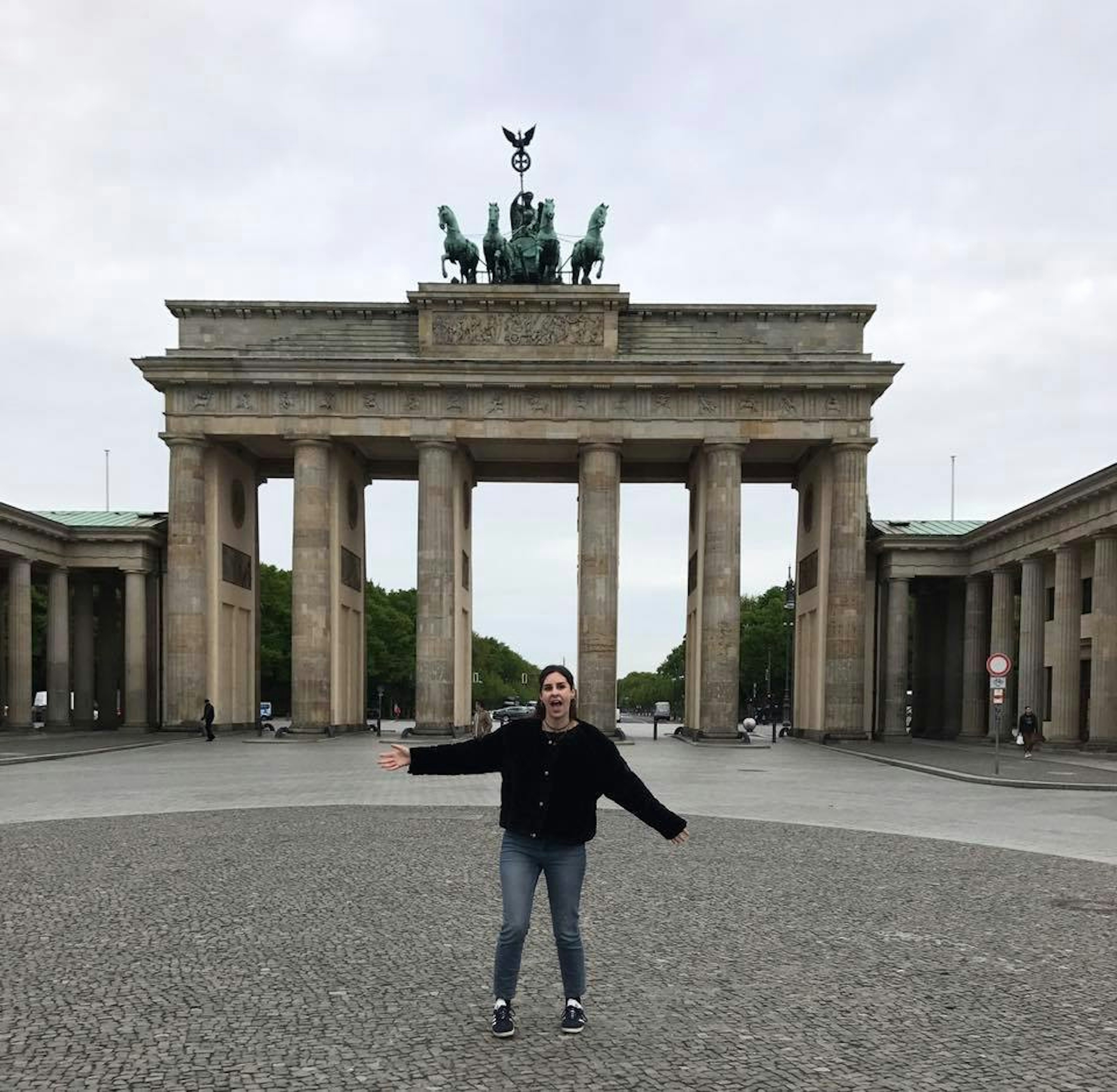 A woman wearing a black jumper and jeans stands with her arms held wide in front of the Brandenburg Gate, a large stone gateway with a statue of horses on top, in Berlin