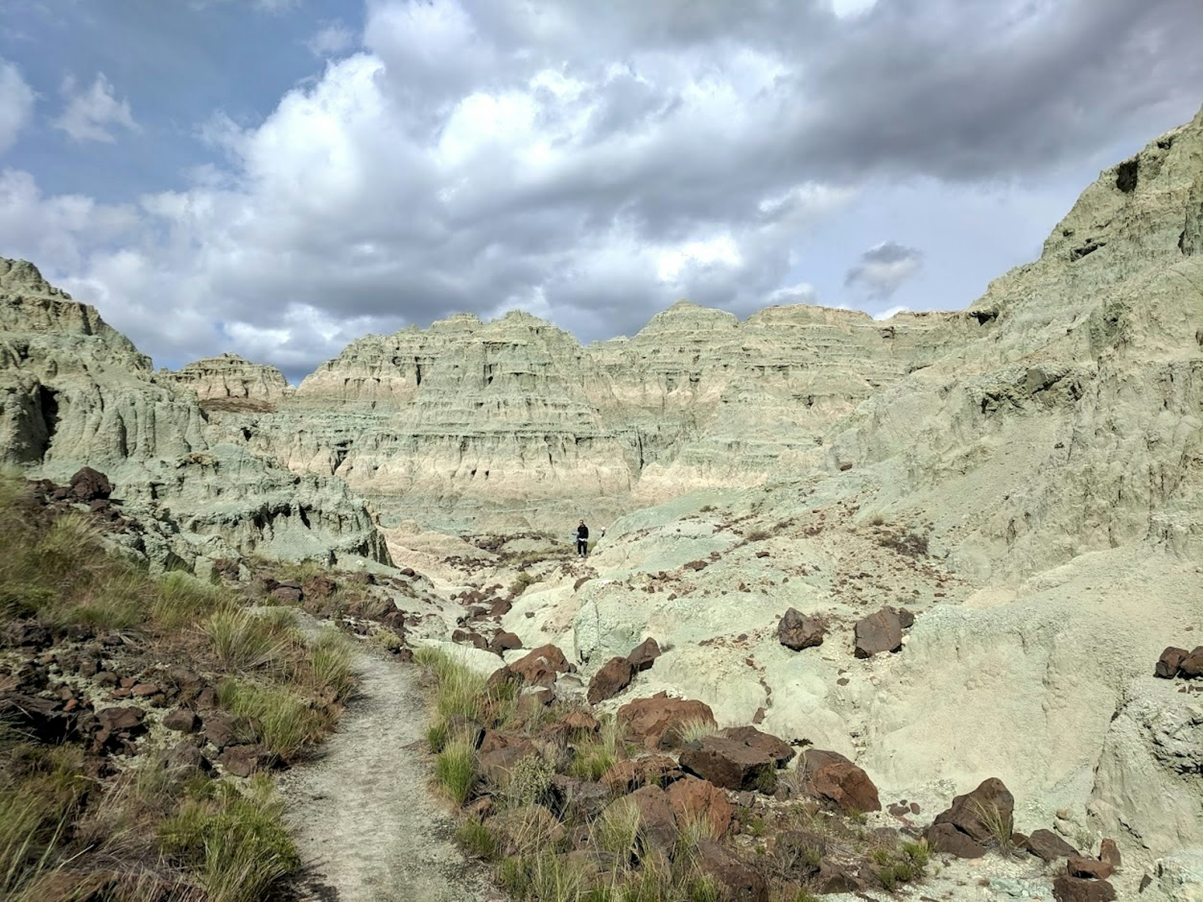 Blue tinted hills in eastern Oregon are cut by a trail at the center of the frame. In the distance, a figure in a black sweat suits walks towards the hills