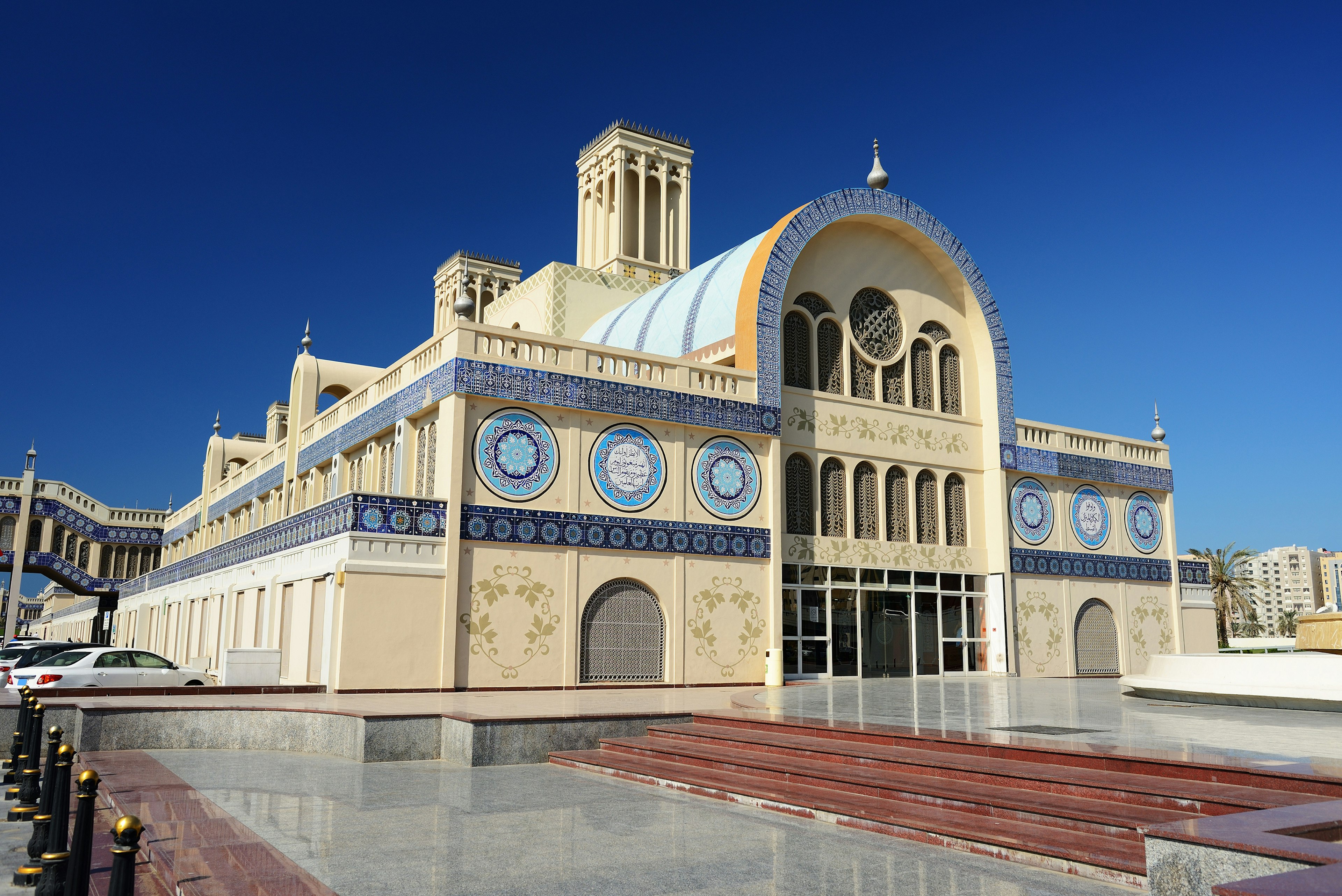 Under a cobalt-blue sky sits a large ornate building with a long cylindrical roof with several square towers; the outside is dominated by a creamy yellow colour, with blue accents.