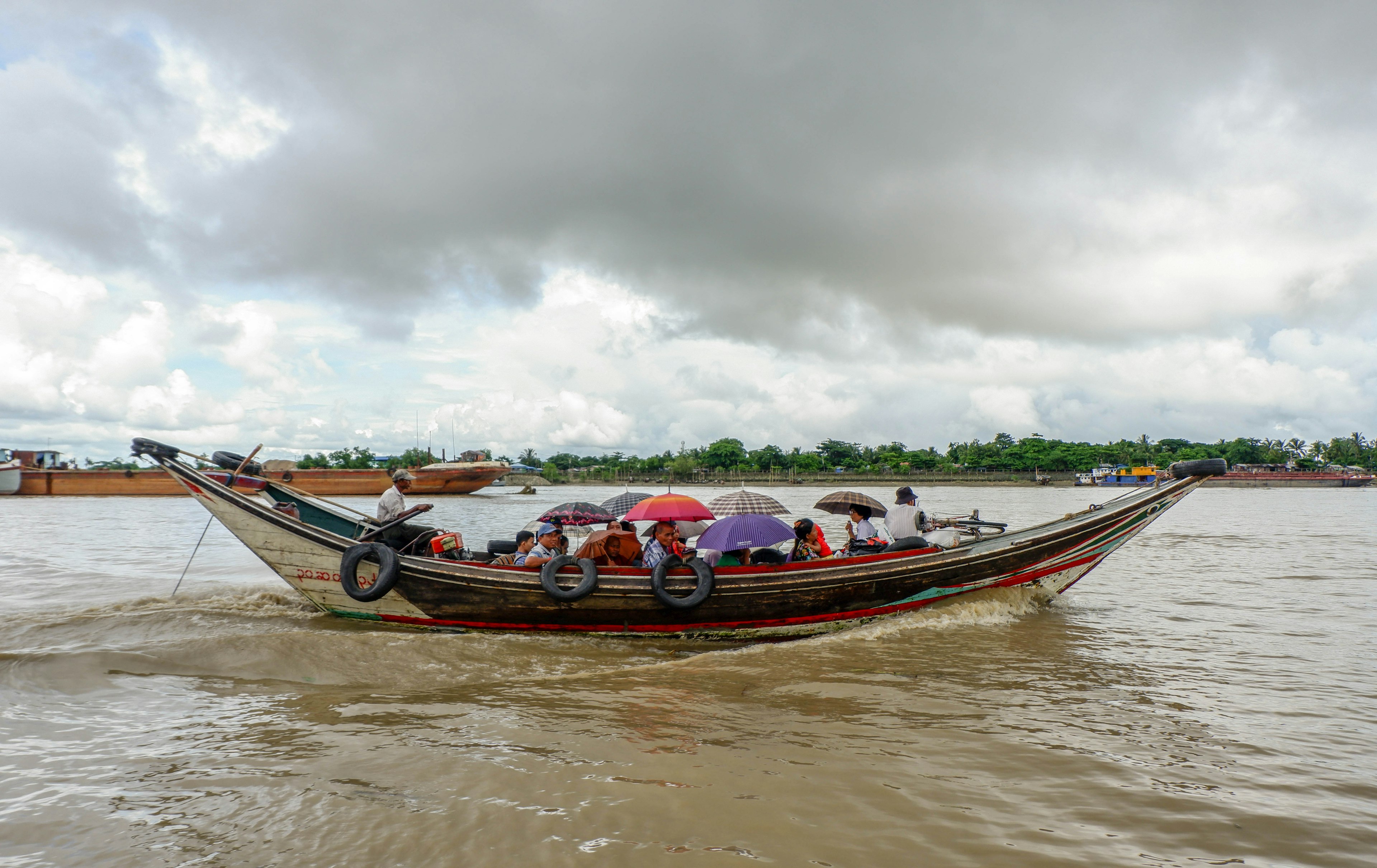 A group of people, some with unbrellas aboard a long, narrow, open boat, are crossing the river towards Seikgyi island.