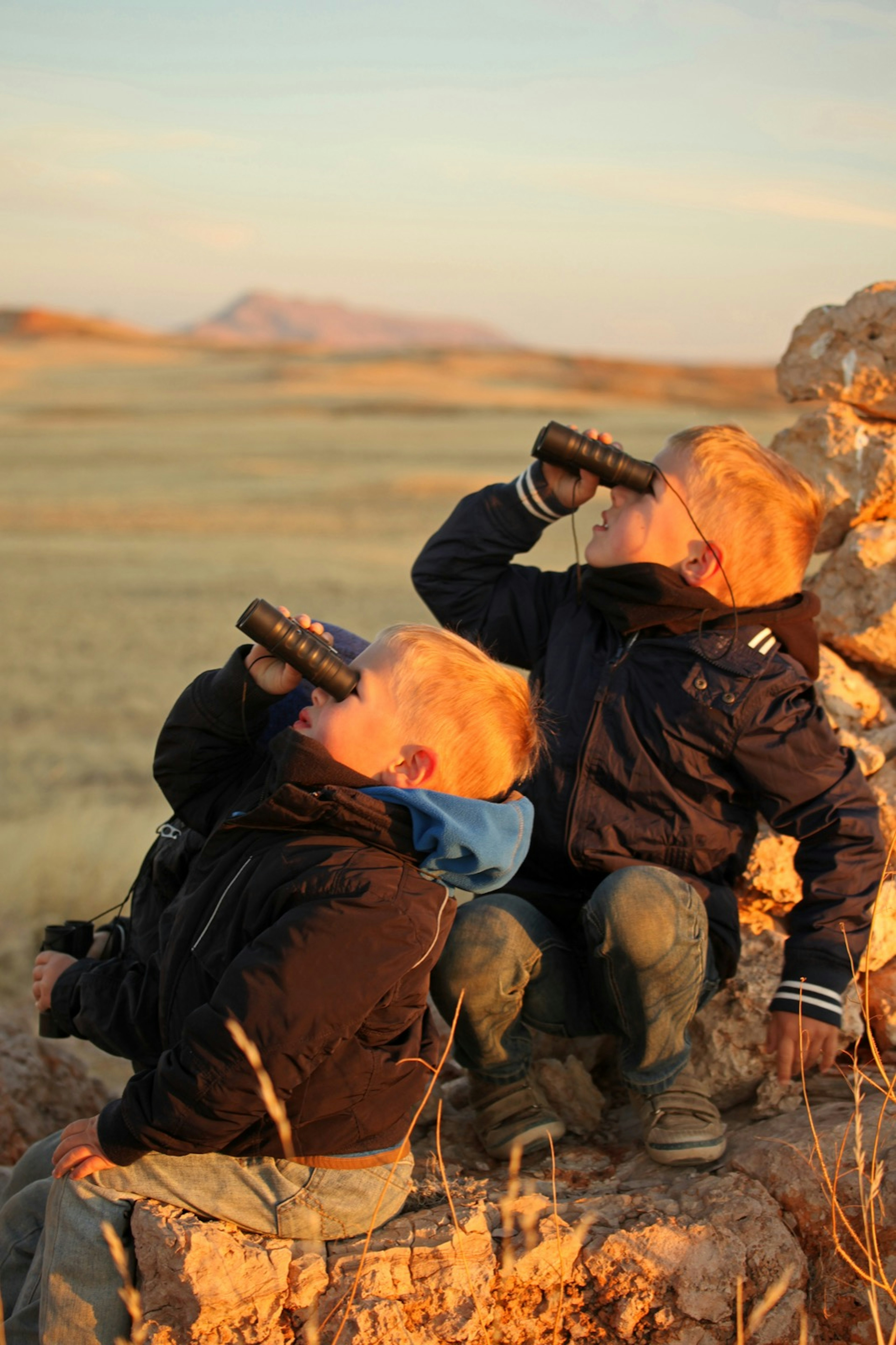 two young boys in black jackets look up to the sky with binoculars