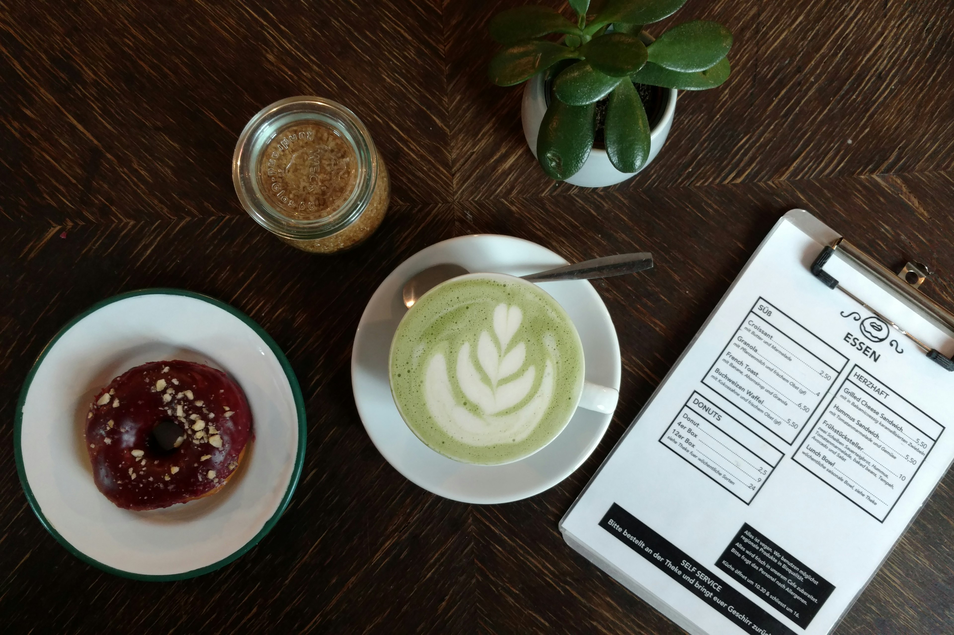 Berlin's best vegan - A chocolate doughnut sprinkled with chopped nuts and a lovely green matcha latte sit next to Brammibal's concise menu and a jar of brown sugar in Berlin.