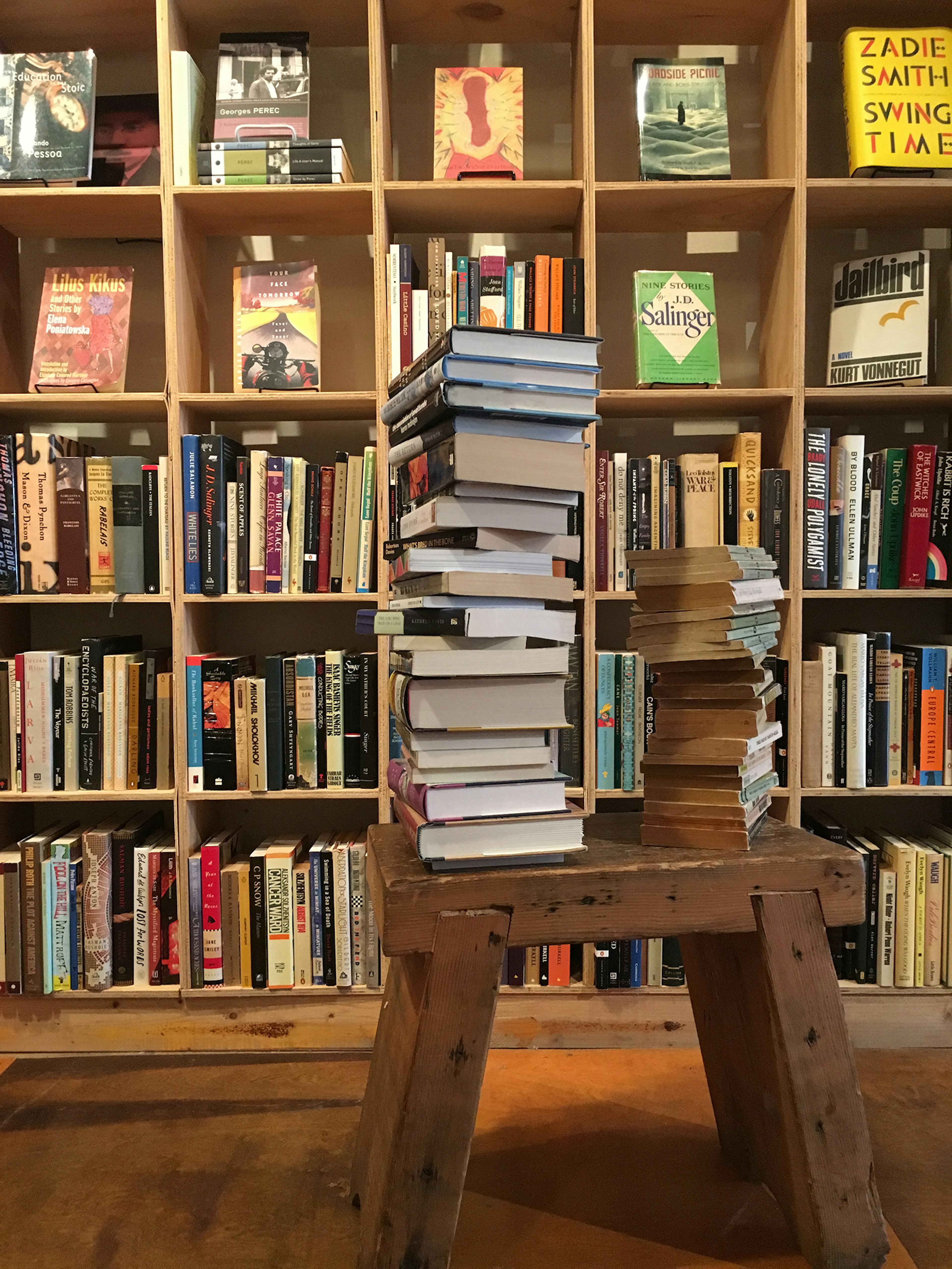 stack of books rests on a wooden bench in front of stocked bookshelves