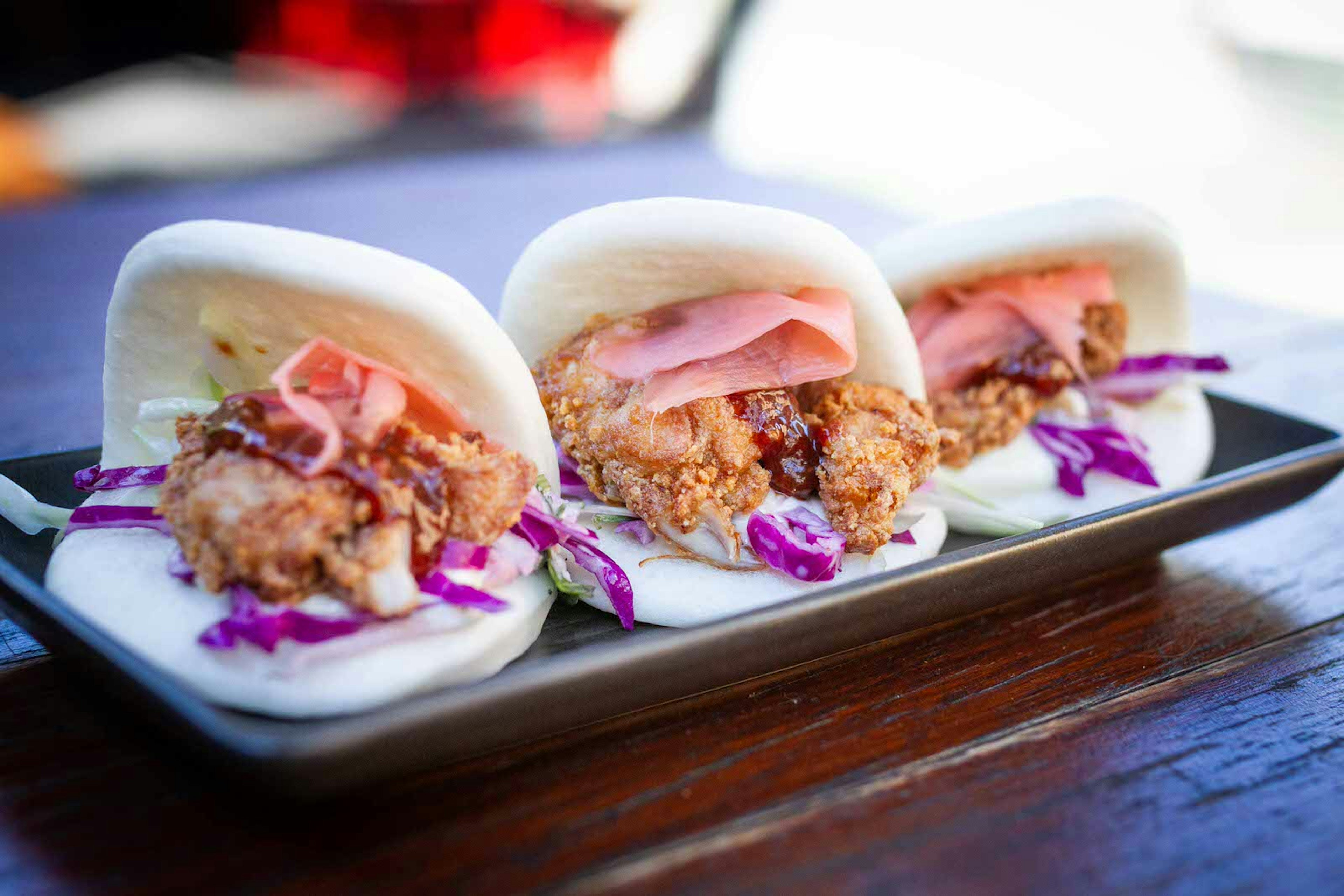 A small rectangular plate on a wood table; on the plate are three identical soft tacos, each filled with deep-fried meat, red cabbage, slices of pickled ginger and bbq sauce.