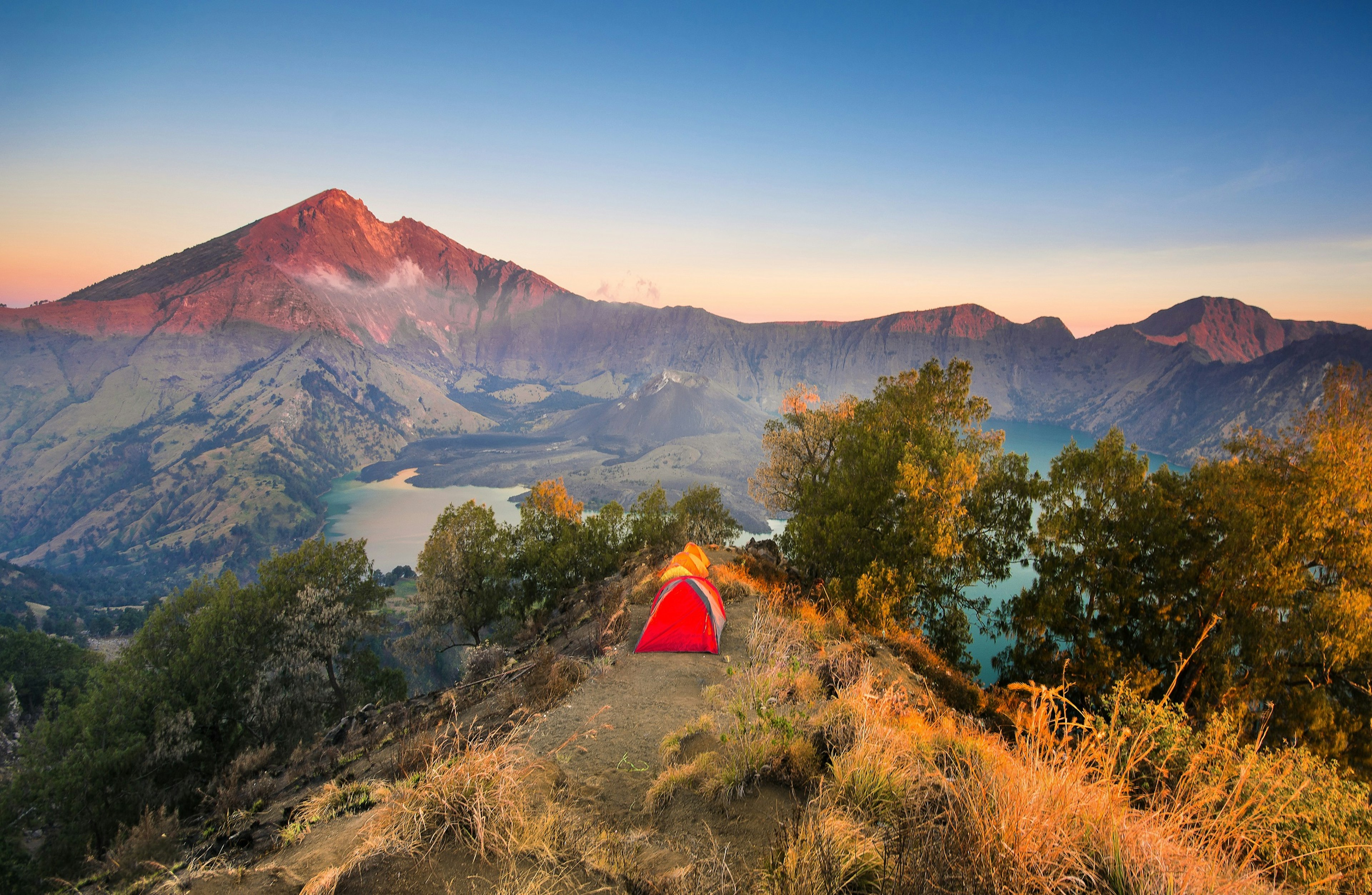 A red and an orange tent sit on a ridge above a lake. A tall peaked mountain is in the background