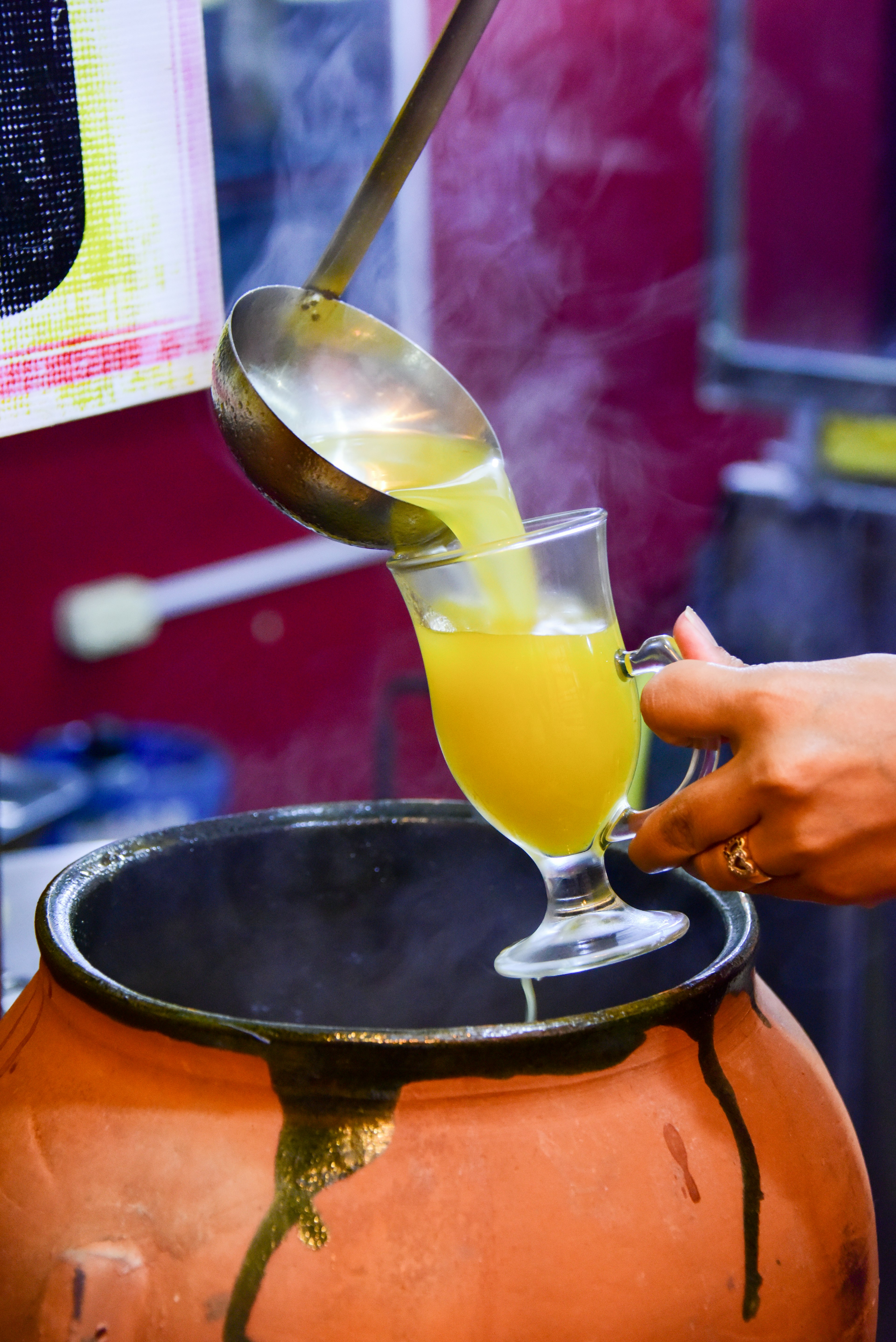 A person ladles canelazo from a brass cauldron into a tall glass