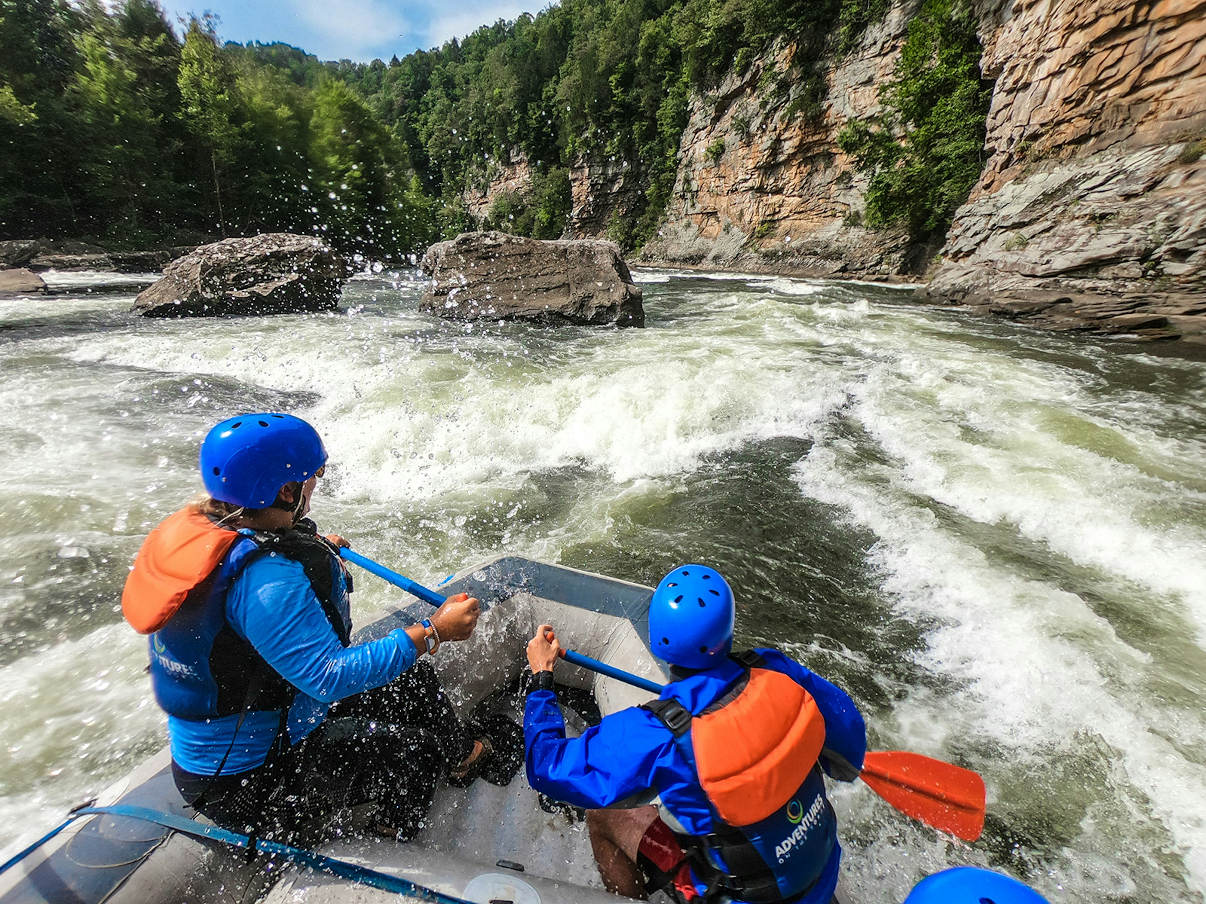 A man and a woman hold paddles at the front of a whitewater raft as they enter a rapid that faces red sandstone cliffs in West Virginia