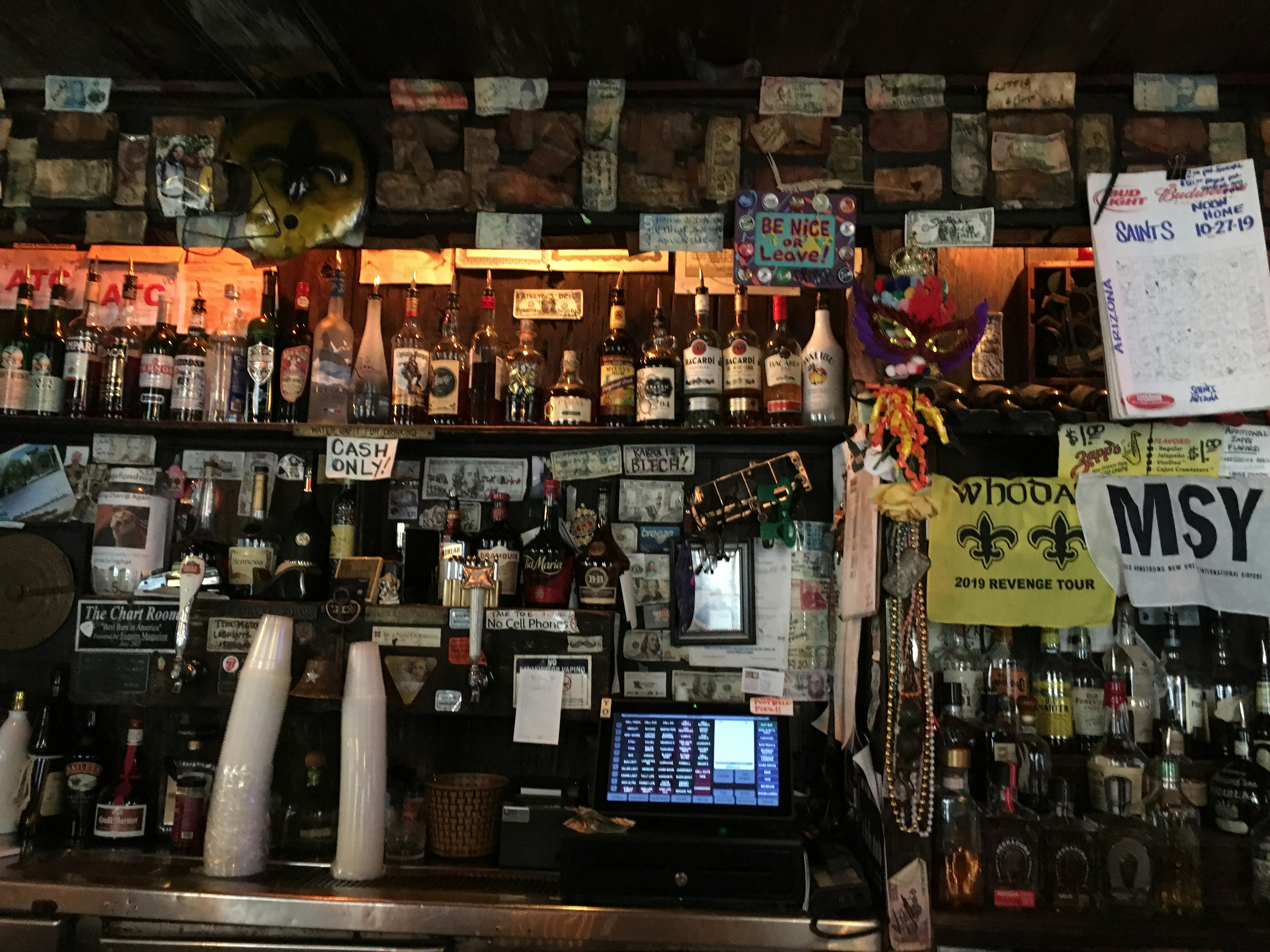 View behind the bar at the Chart Room. Shelved are filled with multiple bottles of alcohol and covered in different currencies, Mardi Gras masks and beads. A cash register is off to the left of a stack of plastic cups.