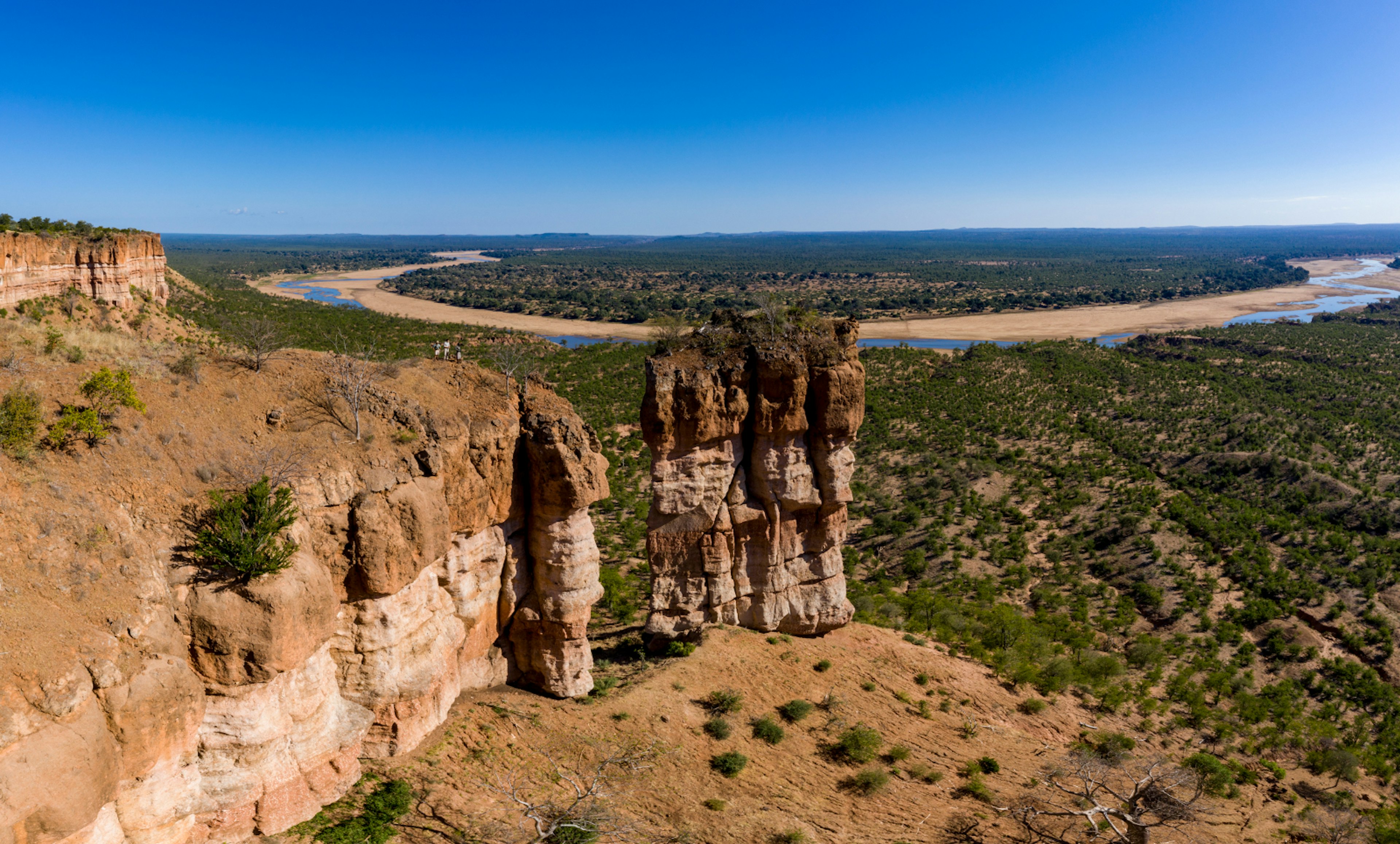 A group of three people stand on the slope between the two-stepped Chilojo Cliffs; in the distance is a huge horseshoe bend in the Runde River.