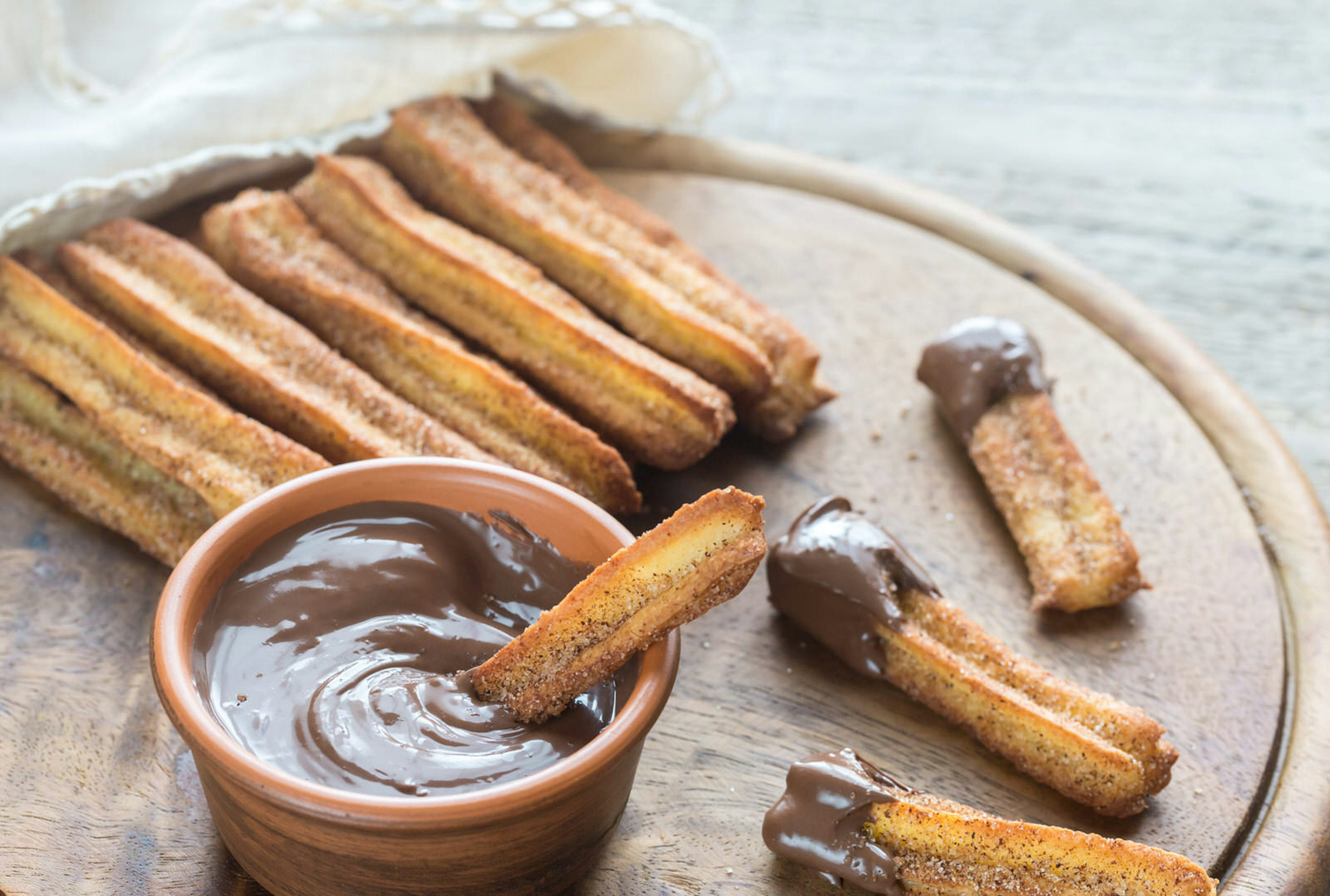 Several churros served on a round, wooden chopping board alongside a terracotta ramekin of chocolate sauce.