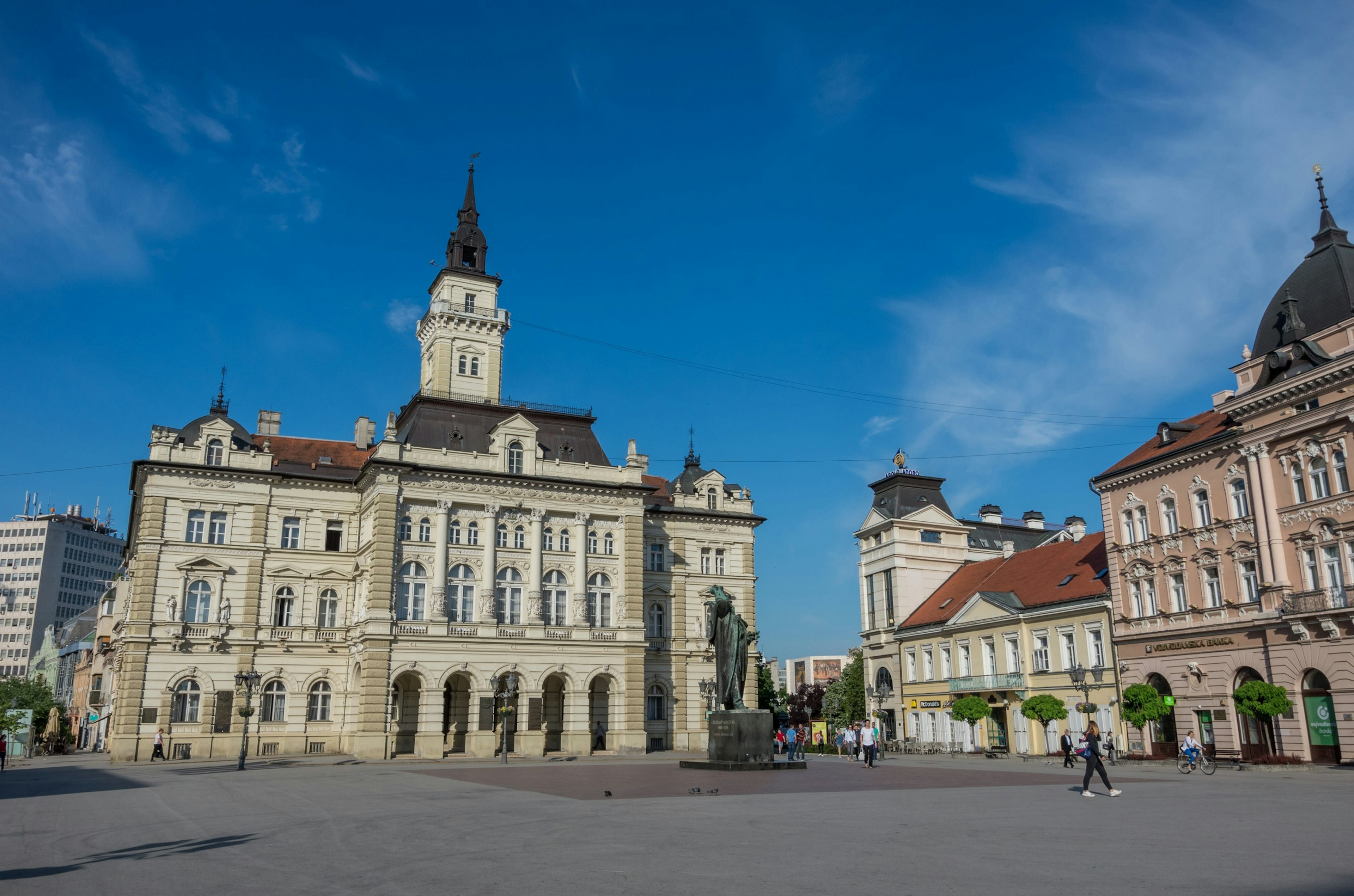 An open square with a large neo-Renaissance-style building to the left. A tall bronze statue of a figure stands in front of the building