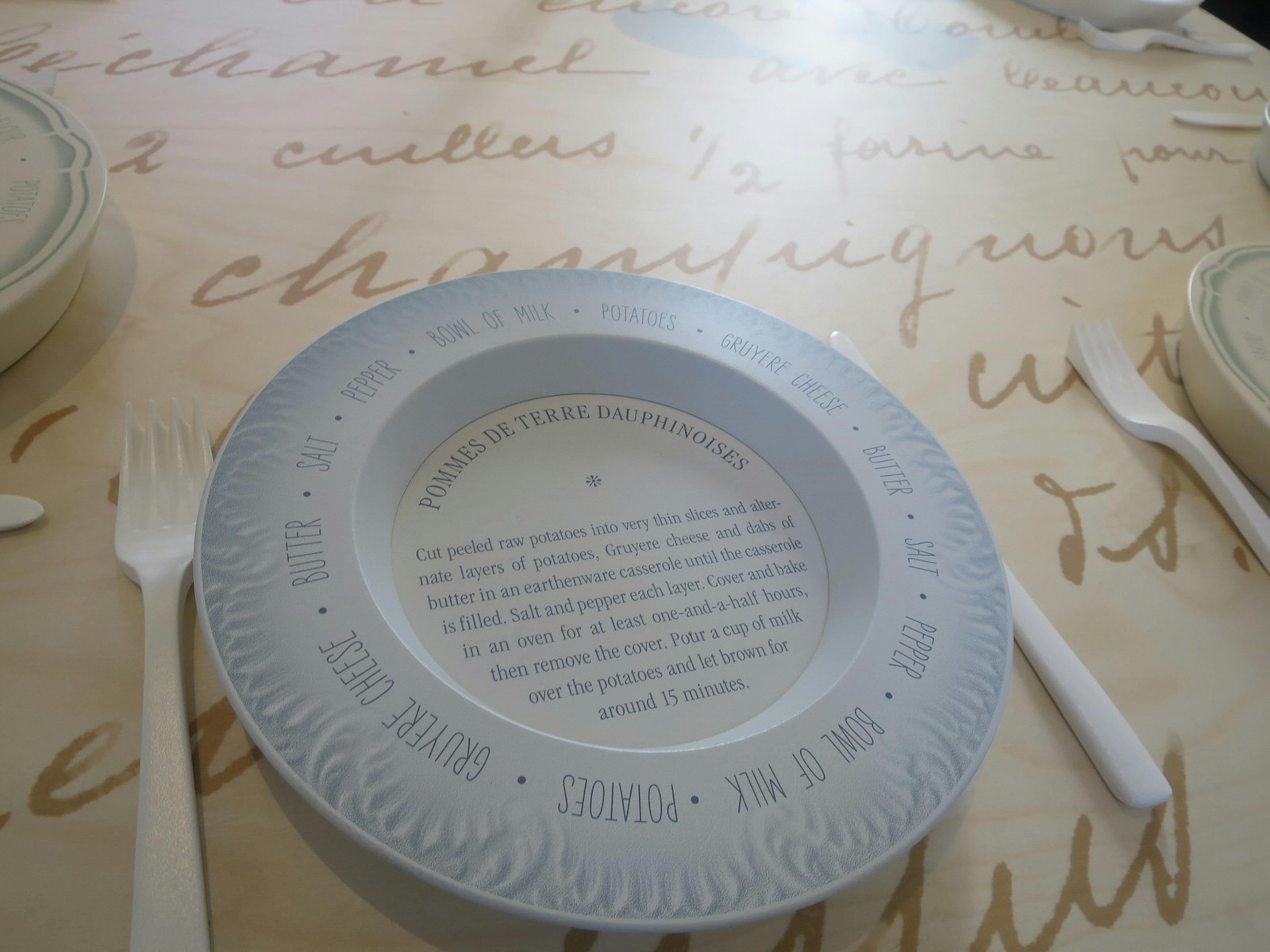 One of the displays of a classic French dishes. A white dish sits on a table with writing on it. The dish has the recipe for 'pommes de terre' written on it.