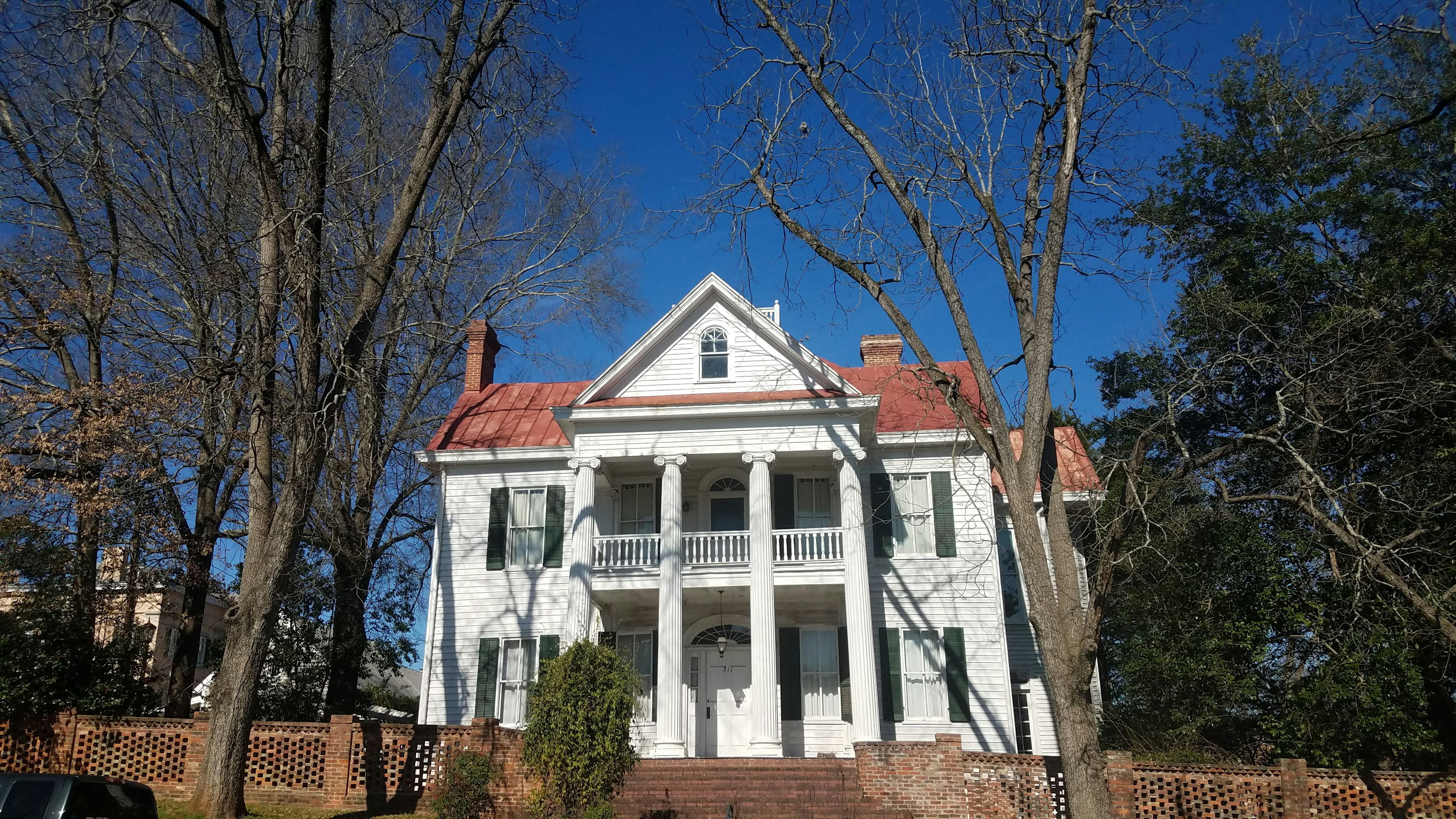 A white antebellum mansion with four Ionic white columns and symmetrical sets of windows on either side of the central porch and a red metal roof sits amidst bare trees behind a low openwork brick wall