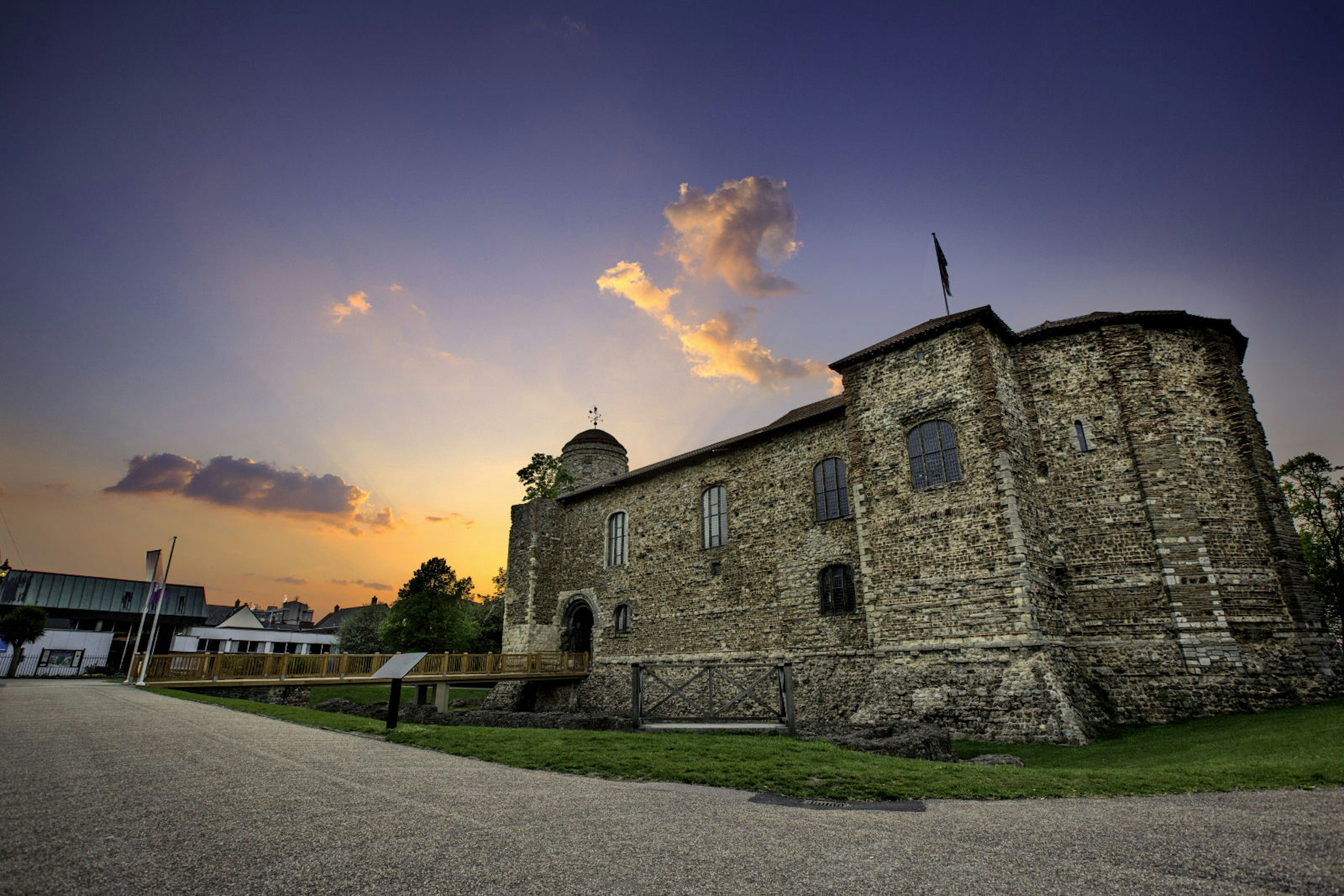 The stone facade of the castle backs against a purple sky with golden light. A modern gravel road is in front of the structure.