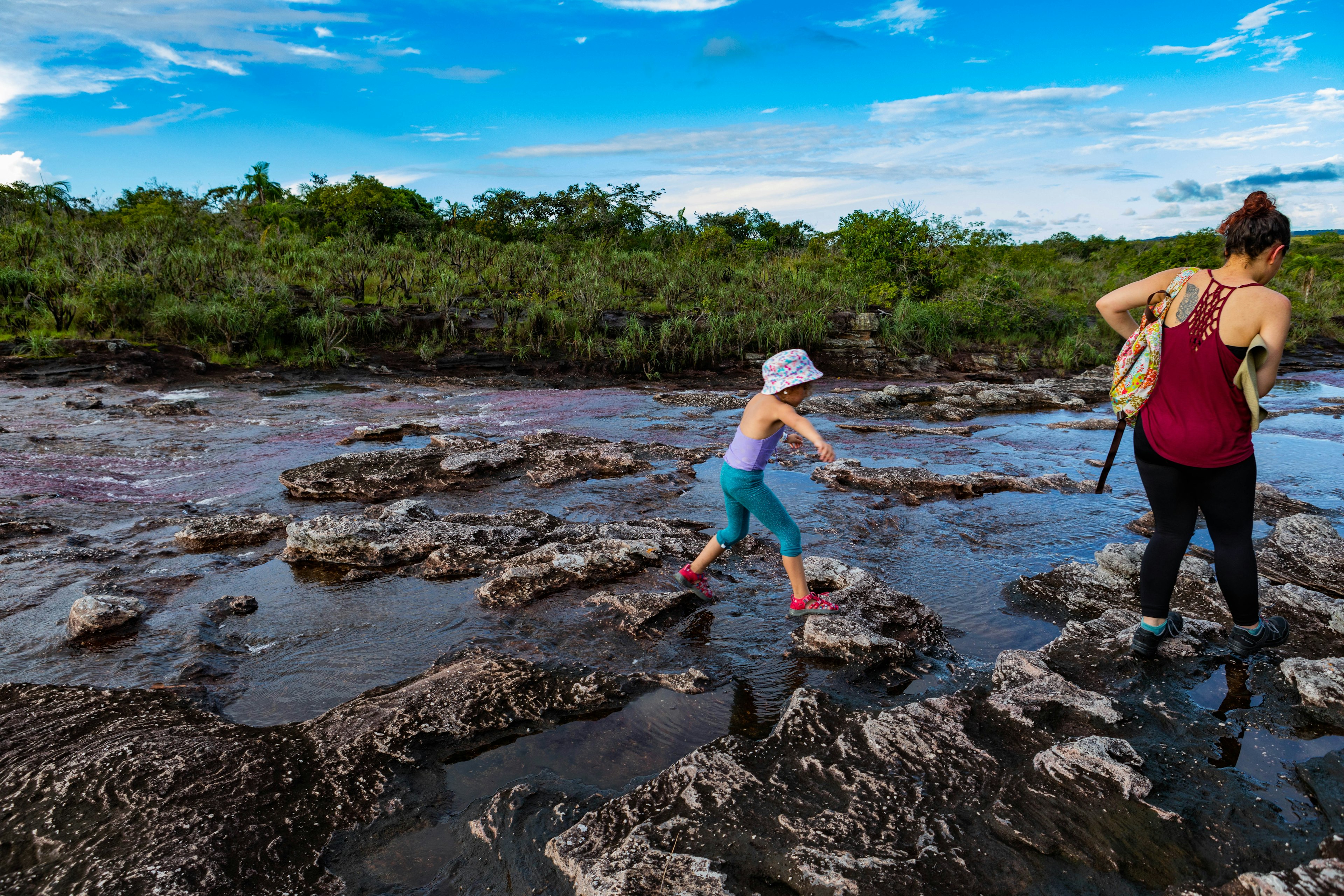 A little girl in blue leggings and a purple swim top hops between rocks on the Cano Cristales river next to a woman in a red tank top and black leggings