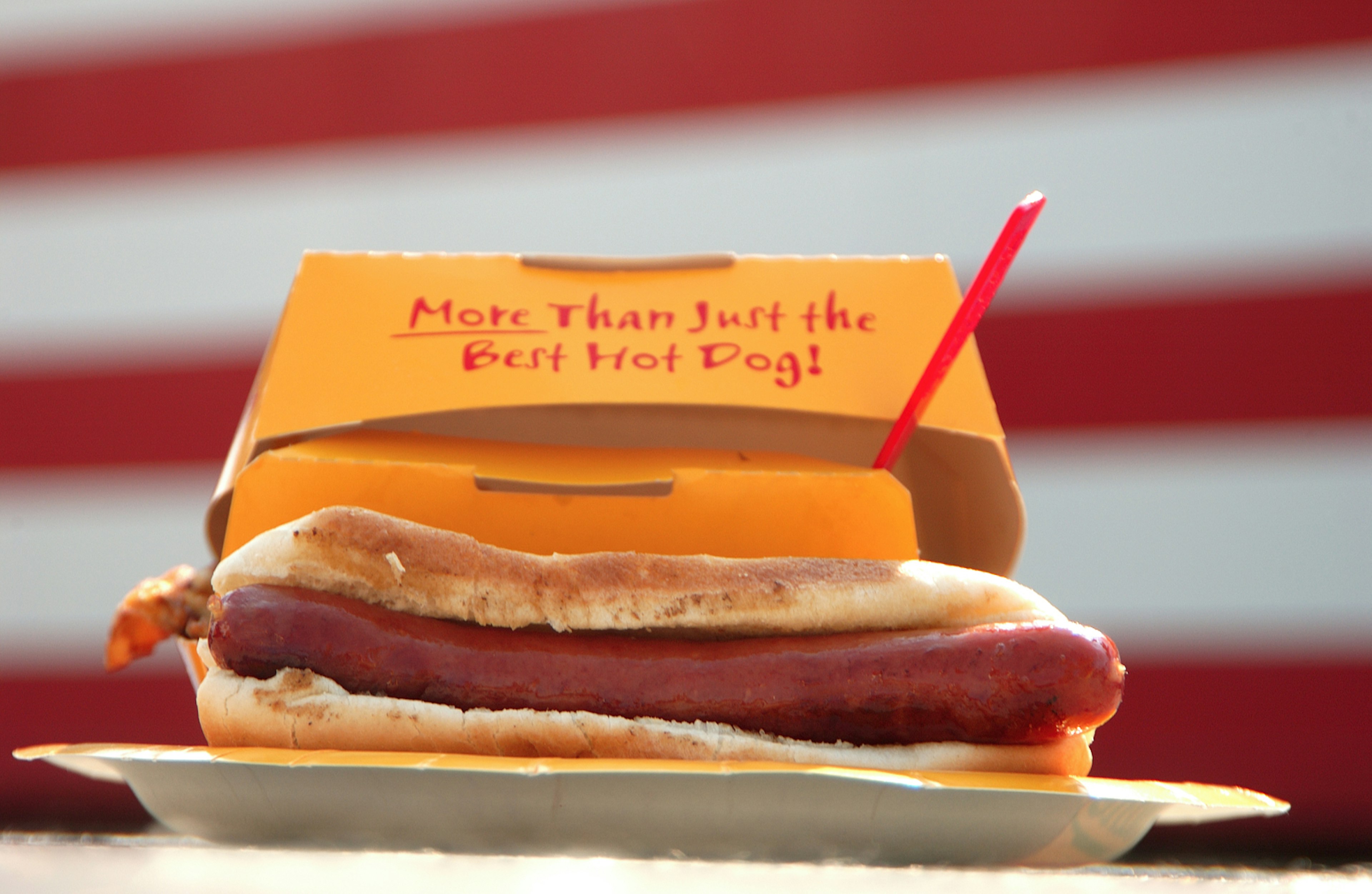 A hot dog in a white bread bun sits on a paper plate, with an orange box behind it that reads: