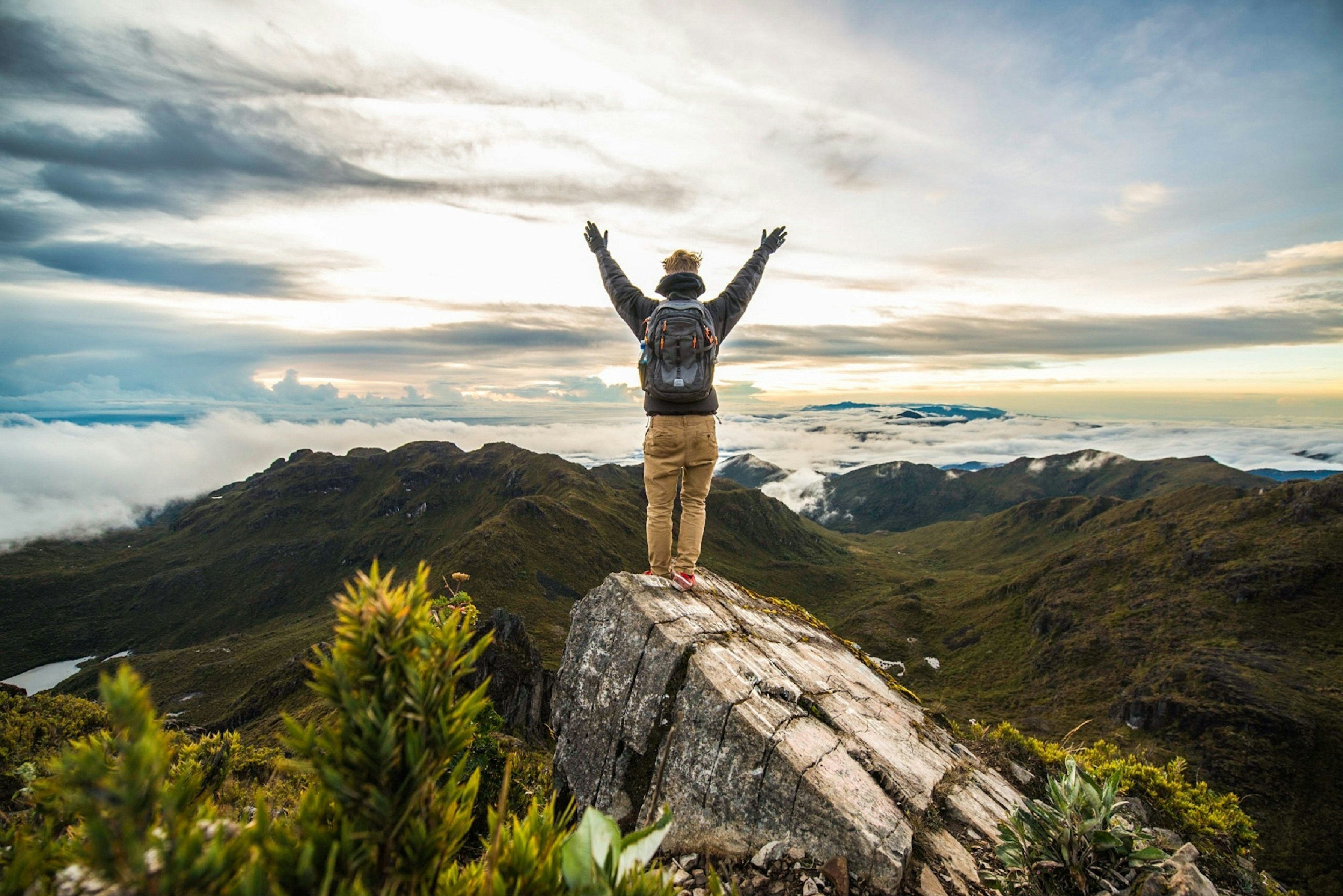 Rear view of man with arms raised standing on rock at Cerro Chirripo, the highest point in the country; below him forested mountains extend into the distance.