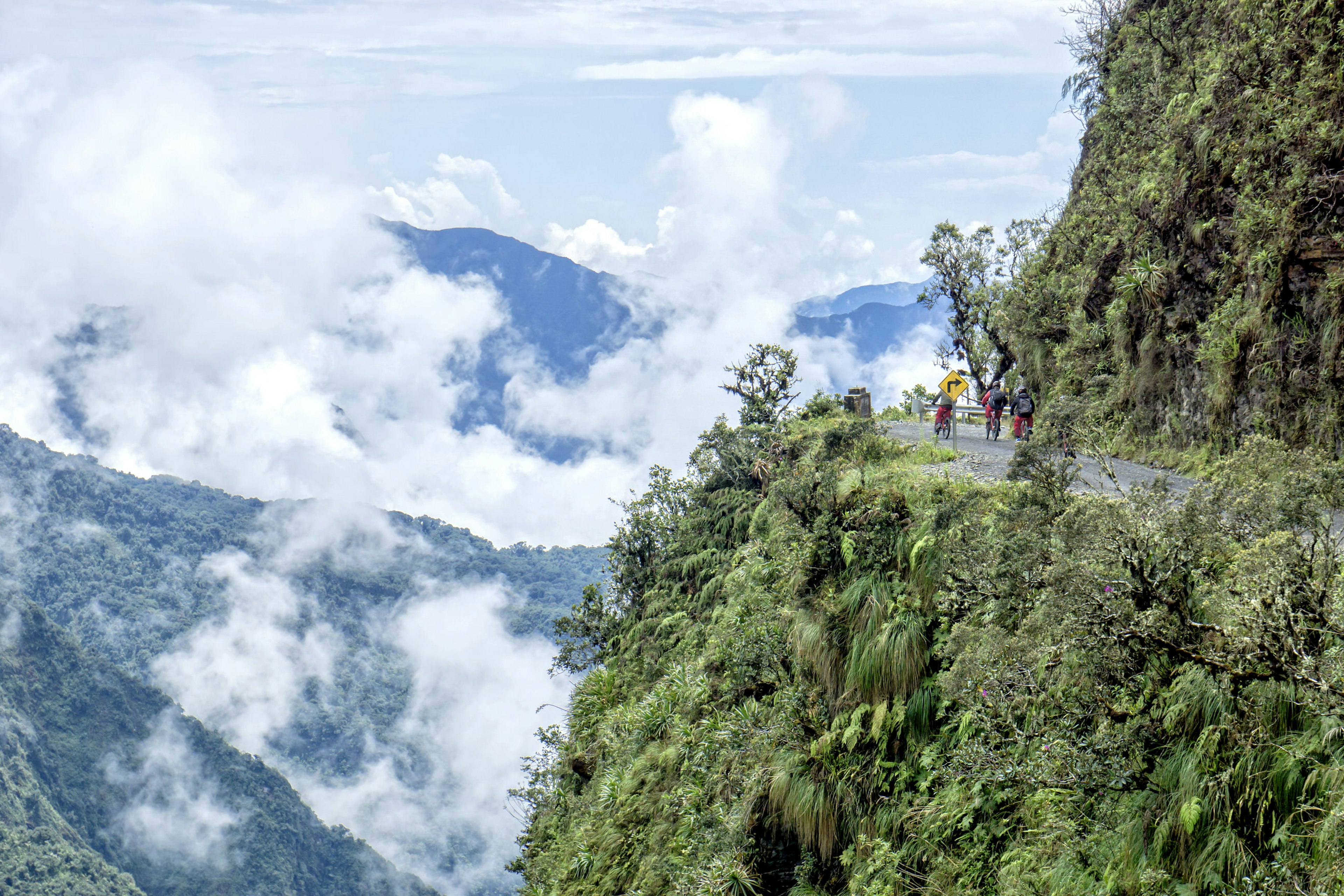 Three mountain bikers ride along a narrow section of road towards a corner; below the road is a 3600m vertical drop down a sheer vegetation-clad cliff. Clouds dot the valley below.