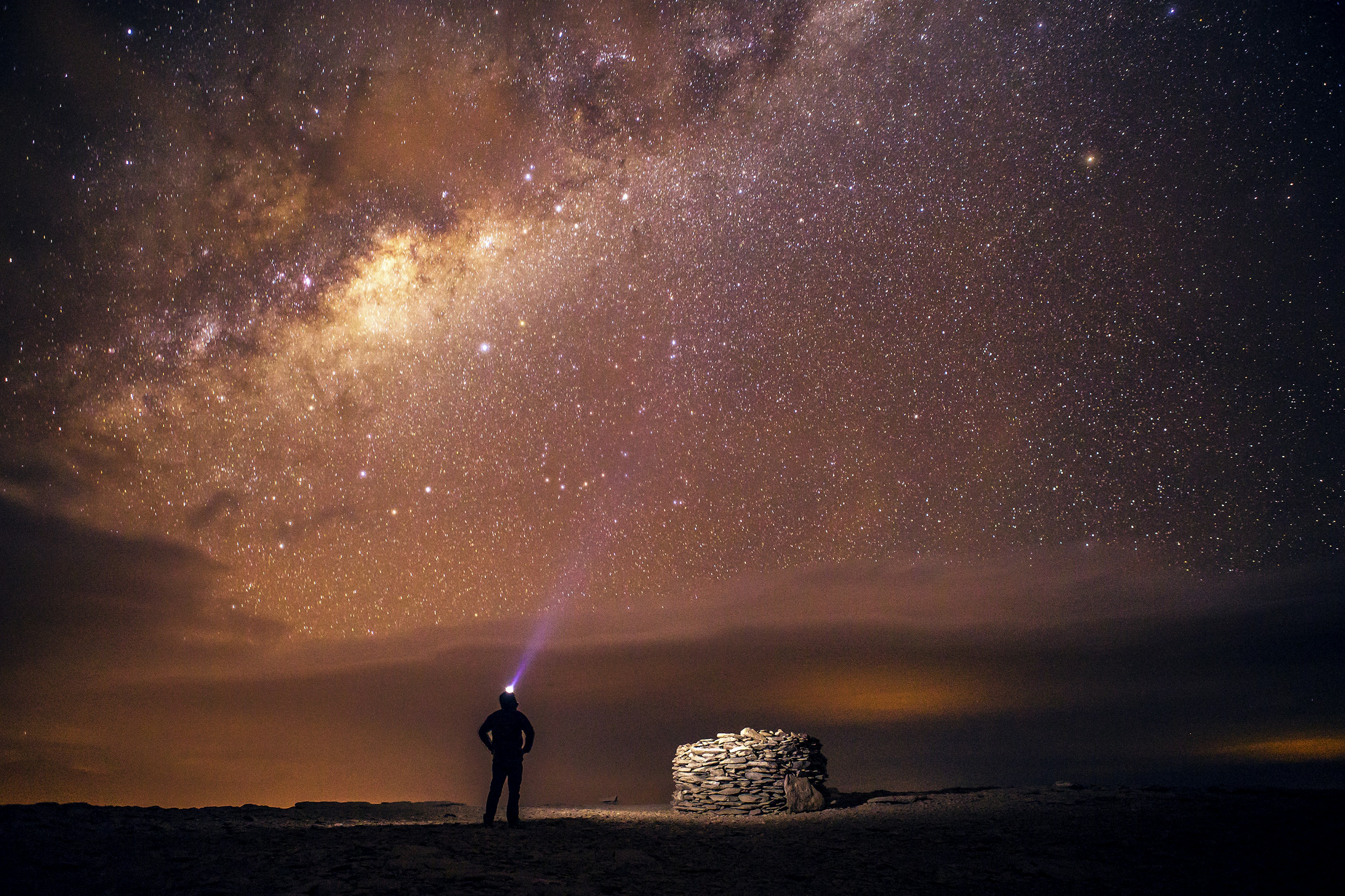 A person stands in the dark next to a stone structure wearing a headlamp; the skies above reveal the Milky Way. Atacama Desert, Chile, South America.