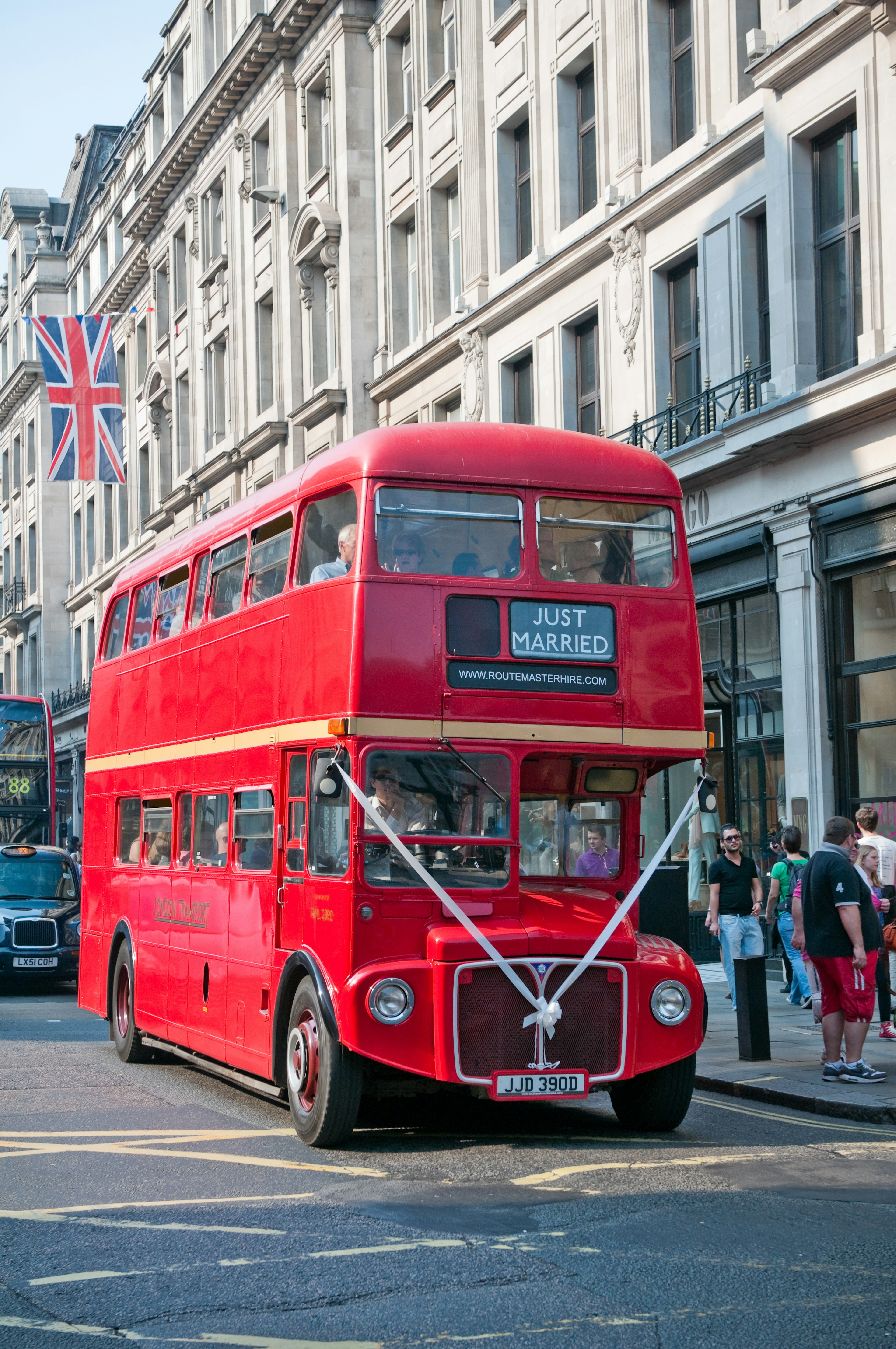 A red Routemaster bus travelling down a London street. It's decorated with white ribbon and bus blind says