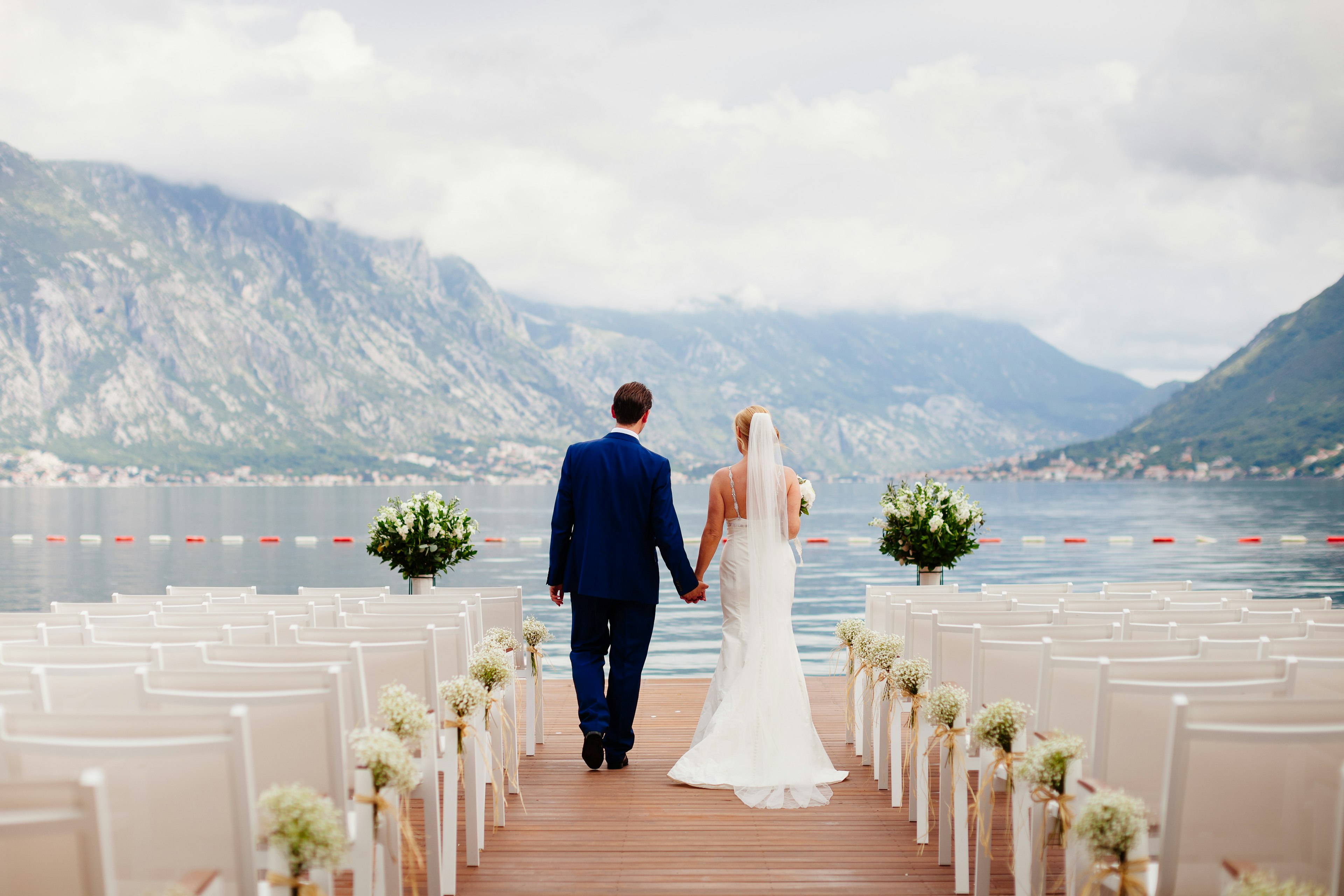 A bride and groom walk between sets of empty chairs laid out for a wedding ceremony in a mountainous lakeside setting.