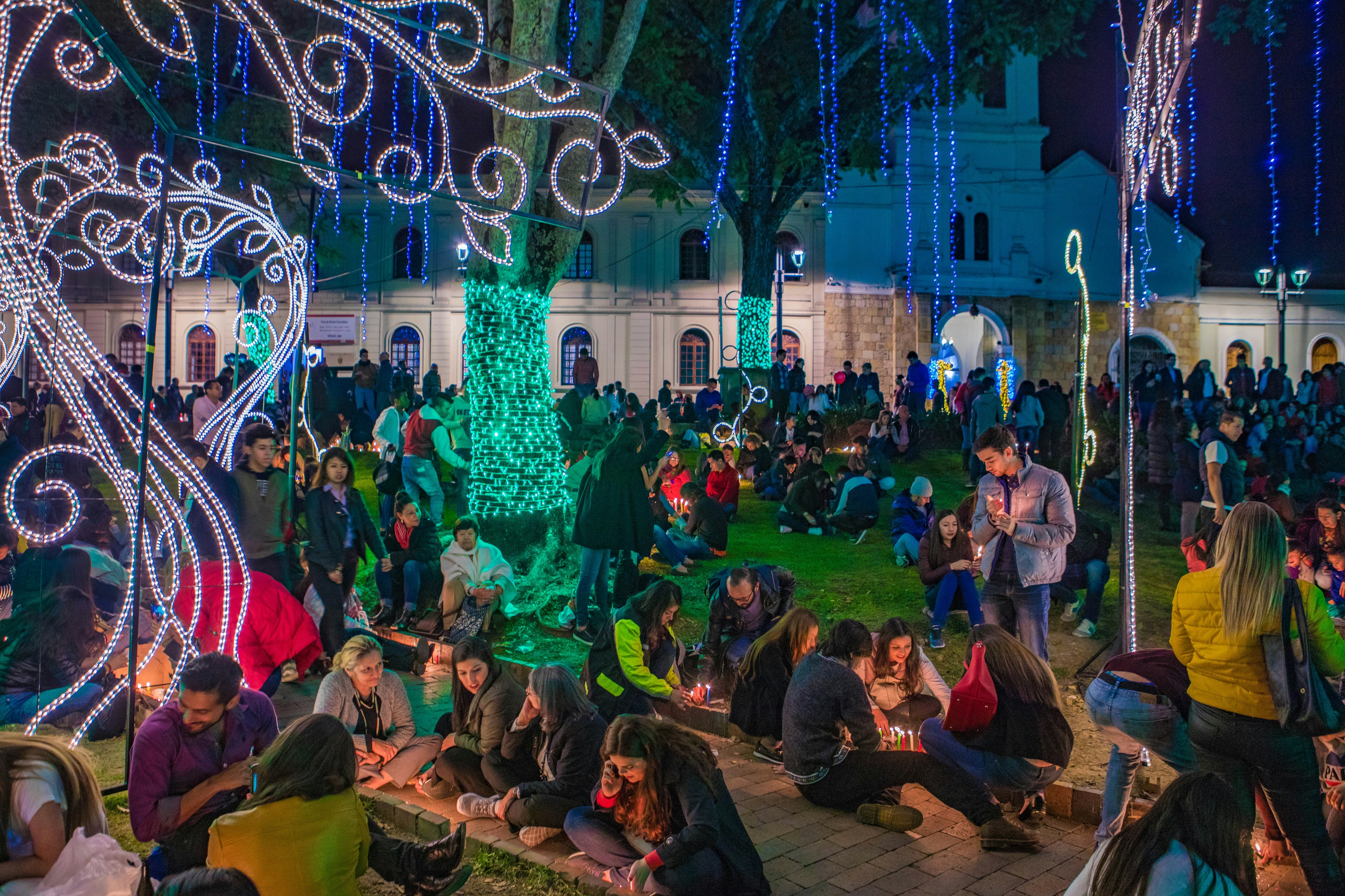 Crowds of people celebrate Dia de Las Velitas by lighting candles and enjoying green Christmas lights wrapped around tree trunks, strands of blue lights dangling from the trees like moss, and white light sculptures in the shape of curlicues and vines standing upright by the sidewalk. In the background are tall white colonial-era stone buildings.