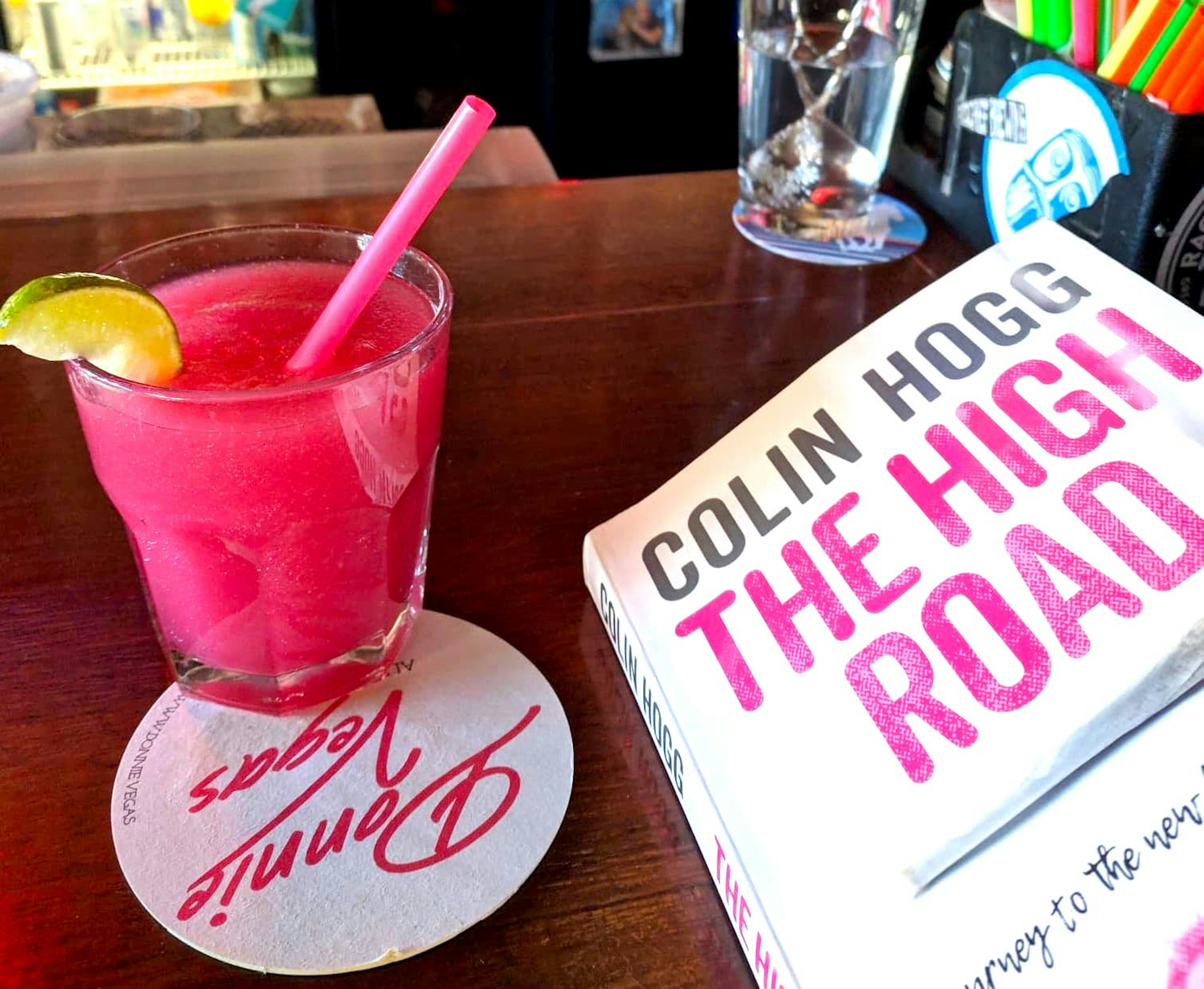 A hot pink cocktail garnished with a lime and with an equally pink plastic straw sits in a highball glass on a coaster reading Donnie Vegas upside down in a fluorescent cursive script. On the right side of the frame is a paperback book by Colin Hogg entitled