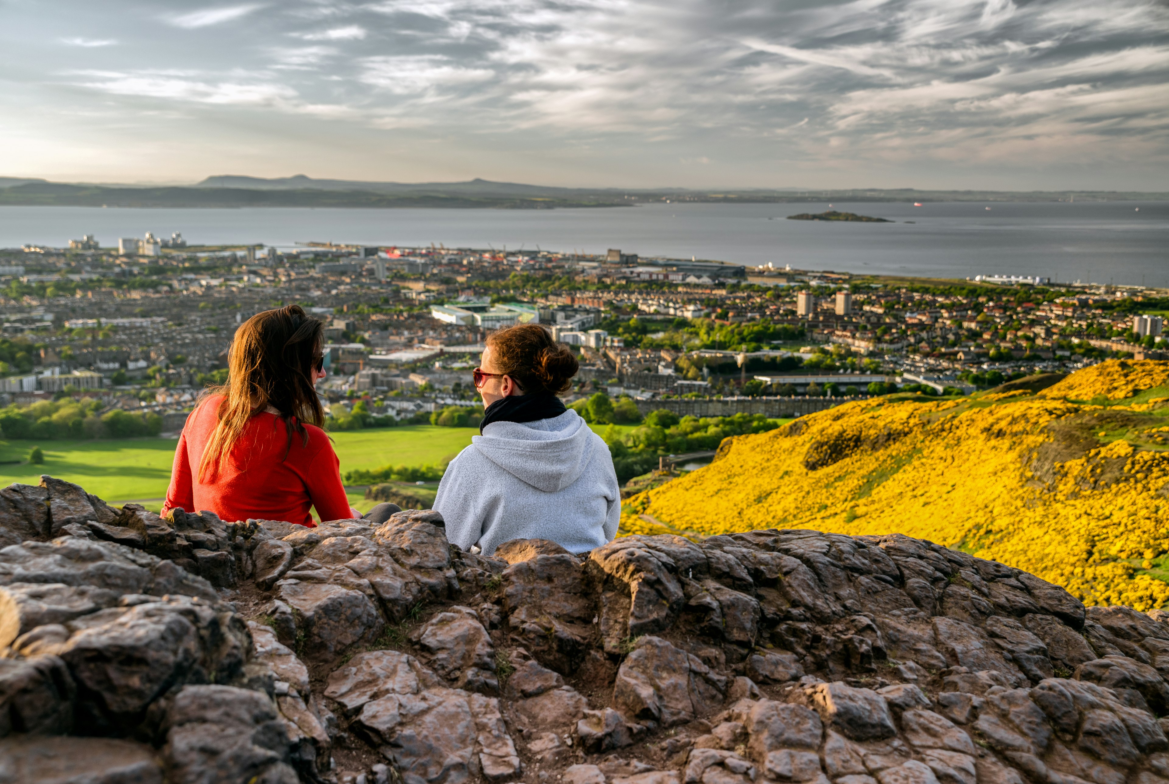 Two girls sitting on the hill of Arthur's Seat overlooking Edinburgh. The hills are covered in yellow wild flowers and the sky is cloudy and moody.