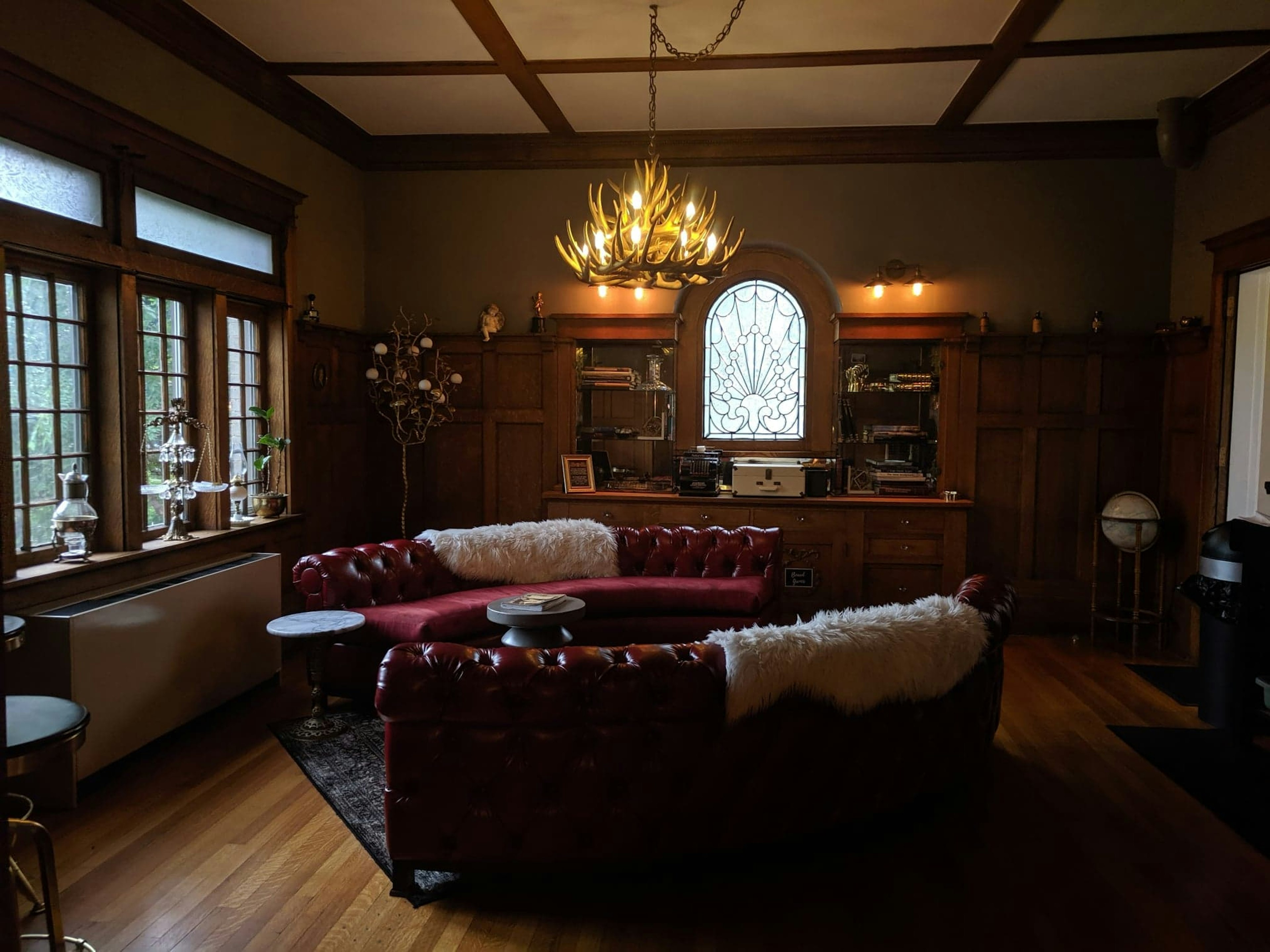 A seating area at Ember Hostel Denver is in an elegant wood paneled room with large glass windows, including one made of clear stained glass surrounded by a built-in credenza. There are two semicircular red leather Chesterfield sofas with white faux sheep skins draped over them. Above is a chandelier made of antlers.