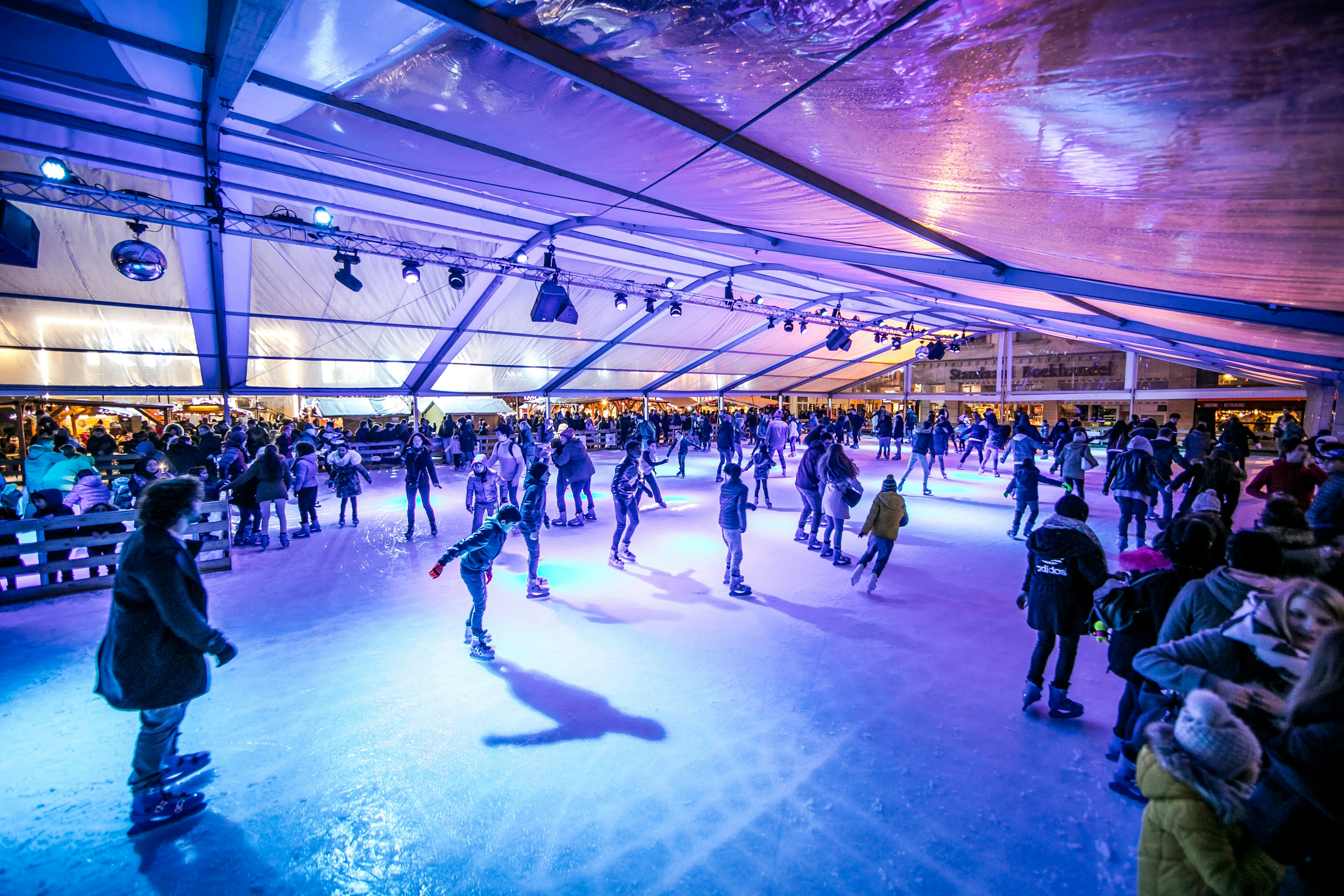 Figure skaters enjoy the rink at Place de la Monnaie, lit with bright purple lighting that makes the ice and the shadows of the skaters glow blue. One young male-presenting figure skater is caught in motion at the center of the image, skating towards his own shadow.