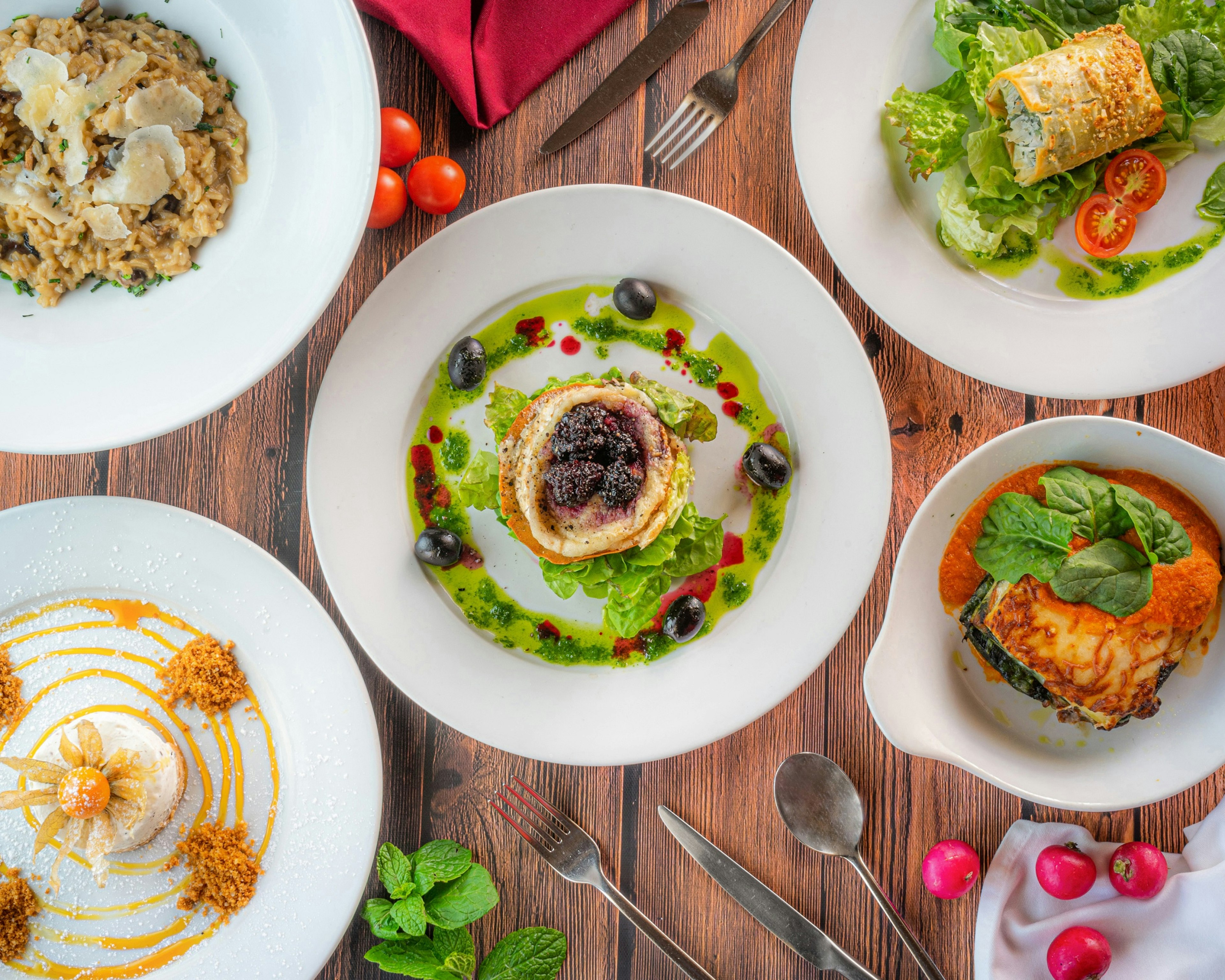 A wooden table showcases five plates of vegetarian fare, some being savoury and others sweet; scattered on the table are some small beets, cherry tomatoes and cutlery.