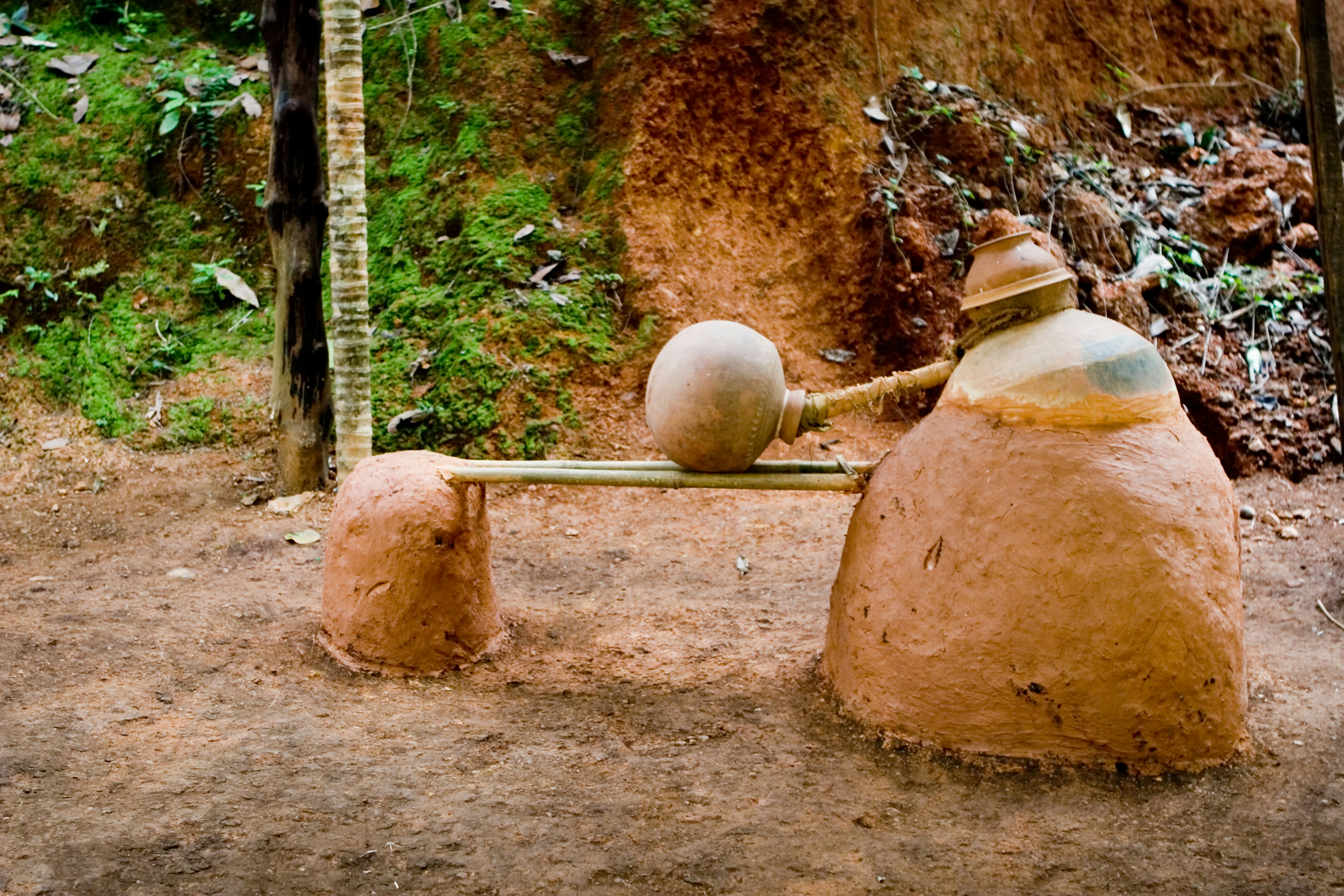 A clay still made up of one large, conical container on the right connected to a rounder container a third of its size on the left, a bar between the two, and a pipe leading to a small spherical container resting on the bar.