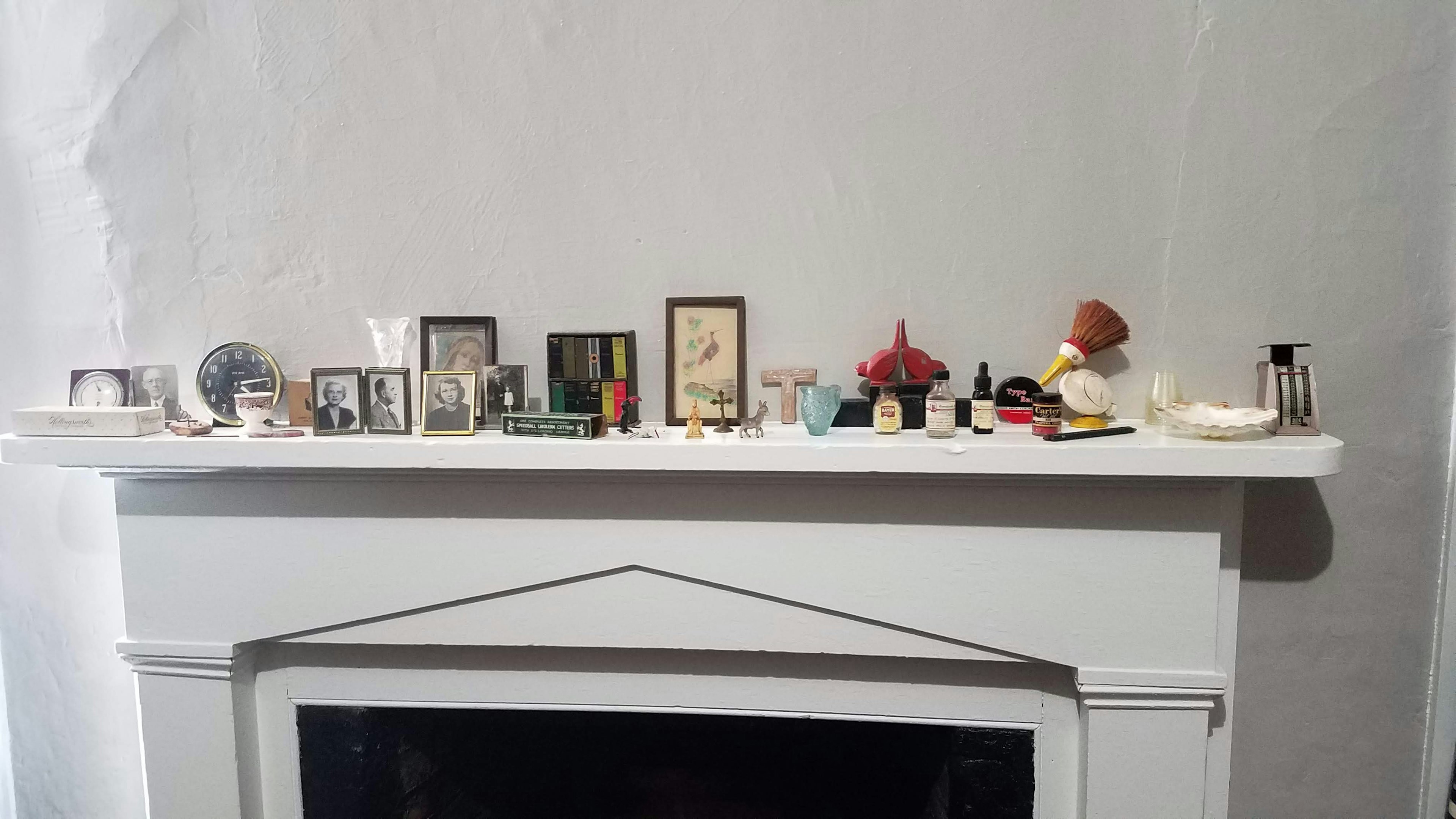 The image shows a white wall with a white mantle covered in framed photos, clocks, and mementos that belonged to Flannery O'Connor in her bedroom at the Andalusia farm in Milledgeville, Georgia