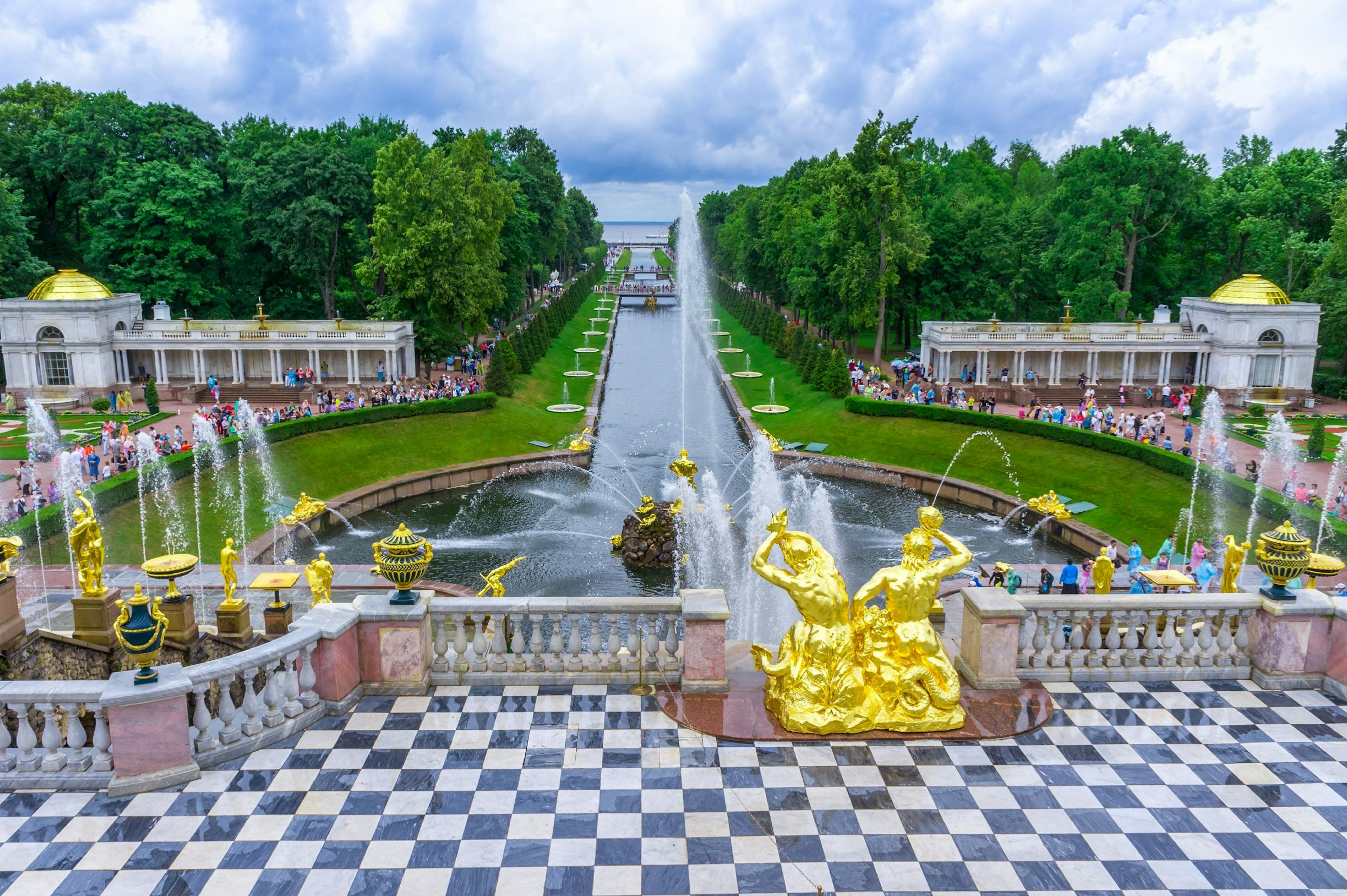 A checked black-and-white tiled terrace with gold statues and fountains beyond. A canal leads to a river, and crowds of people can be seen milling around the water features